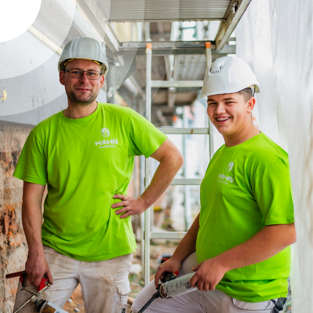 Zwei Menschen in passenden grünen Hemden umarmen sich glücklich in einer belebten Inneneinrichtung, umgeben von anderen, die angeregt über ein spannendes Bauvorhaben diskutieren.