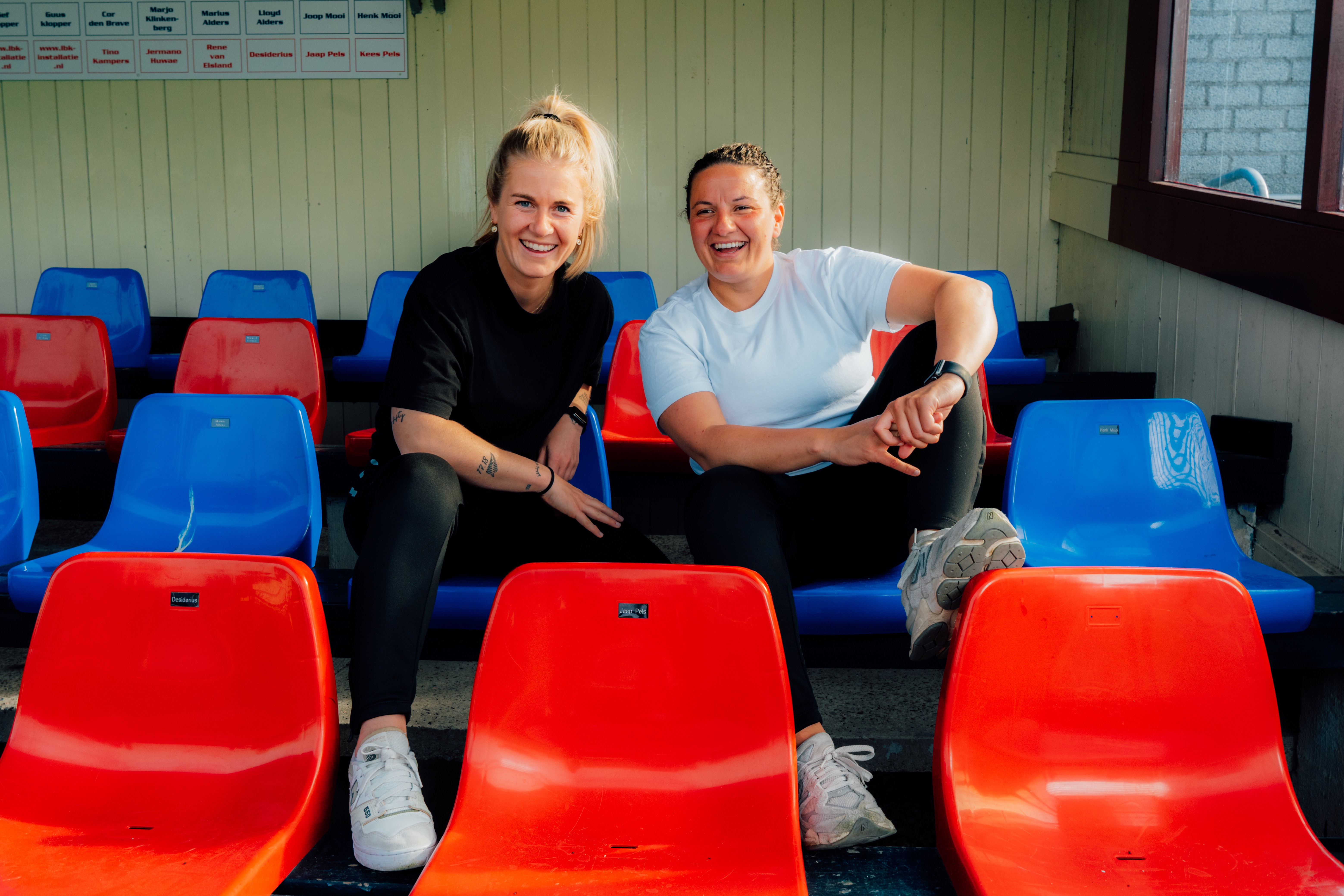 Twee lachende vrouwen zitten ontspannen op rode en blauwe stoelen in een sporttribune.