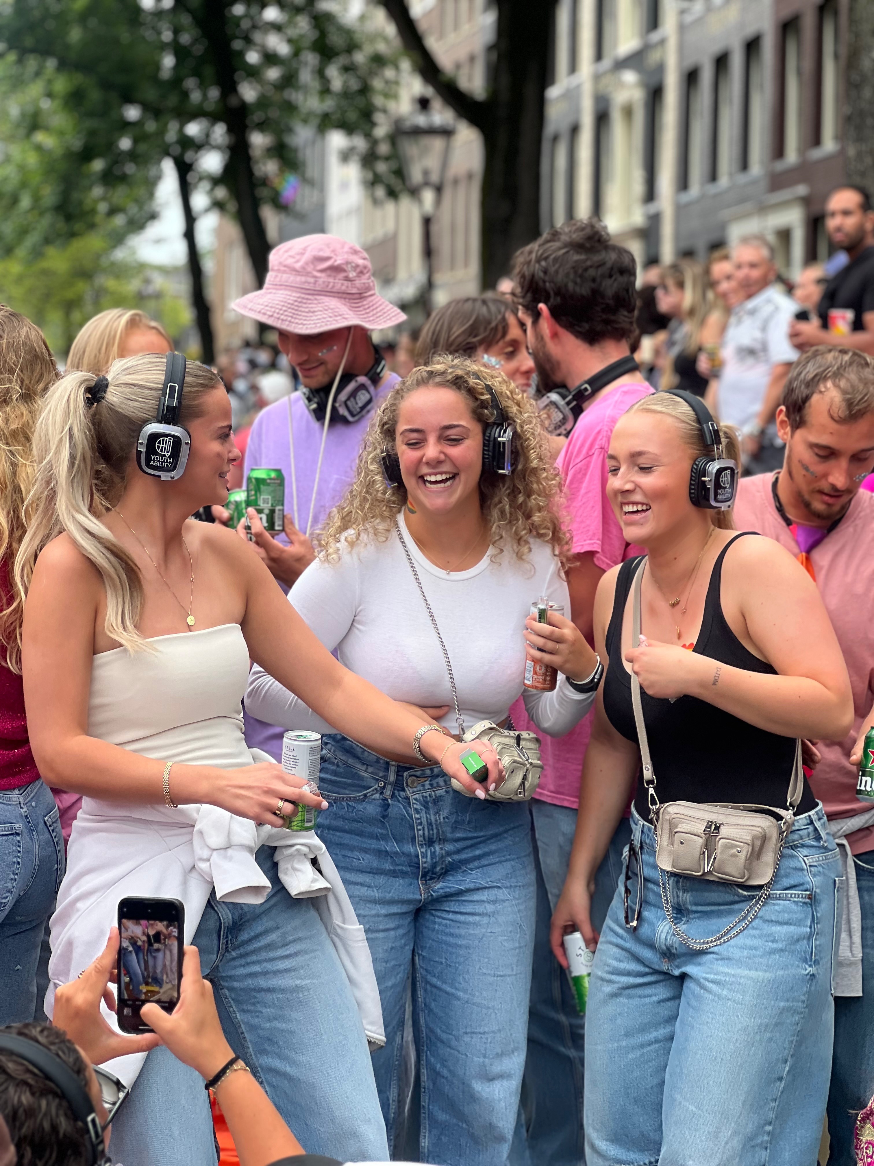 Drie lachende jonge vrouwen met koptelefoons staan buiten op een straatfeest met achter hen een groep feestvierders.