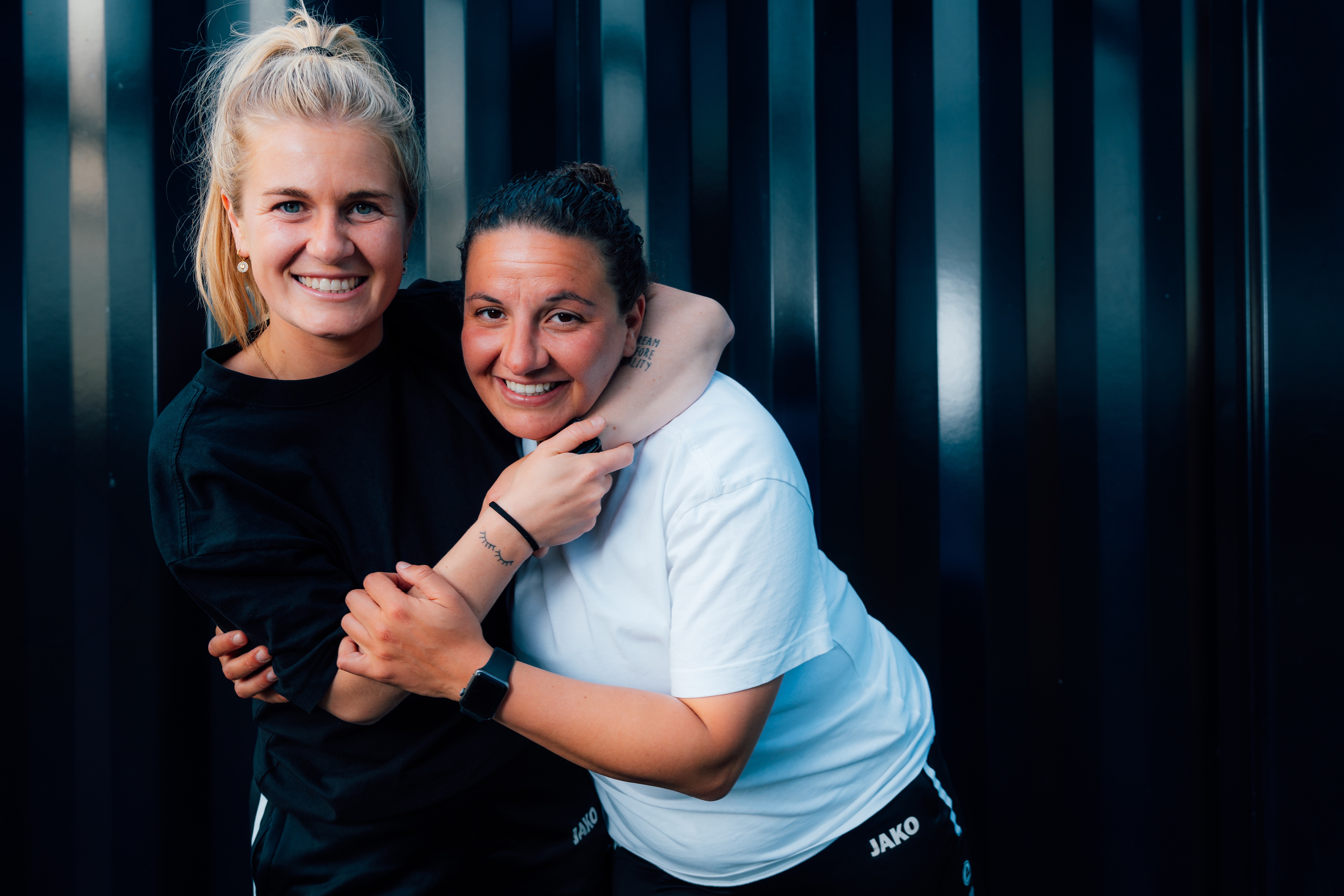Twee vrouwen in sportkleding omhelzen elkaar glimlachend voor een donkere achtergrond.