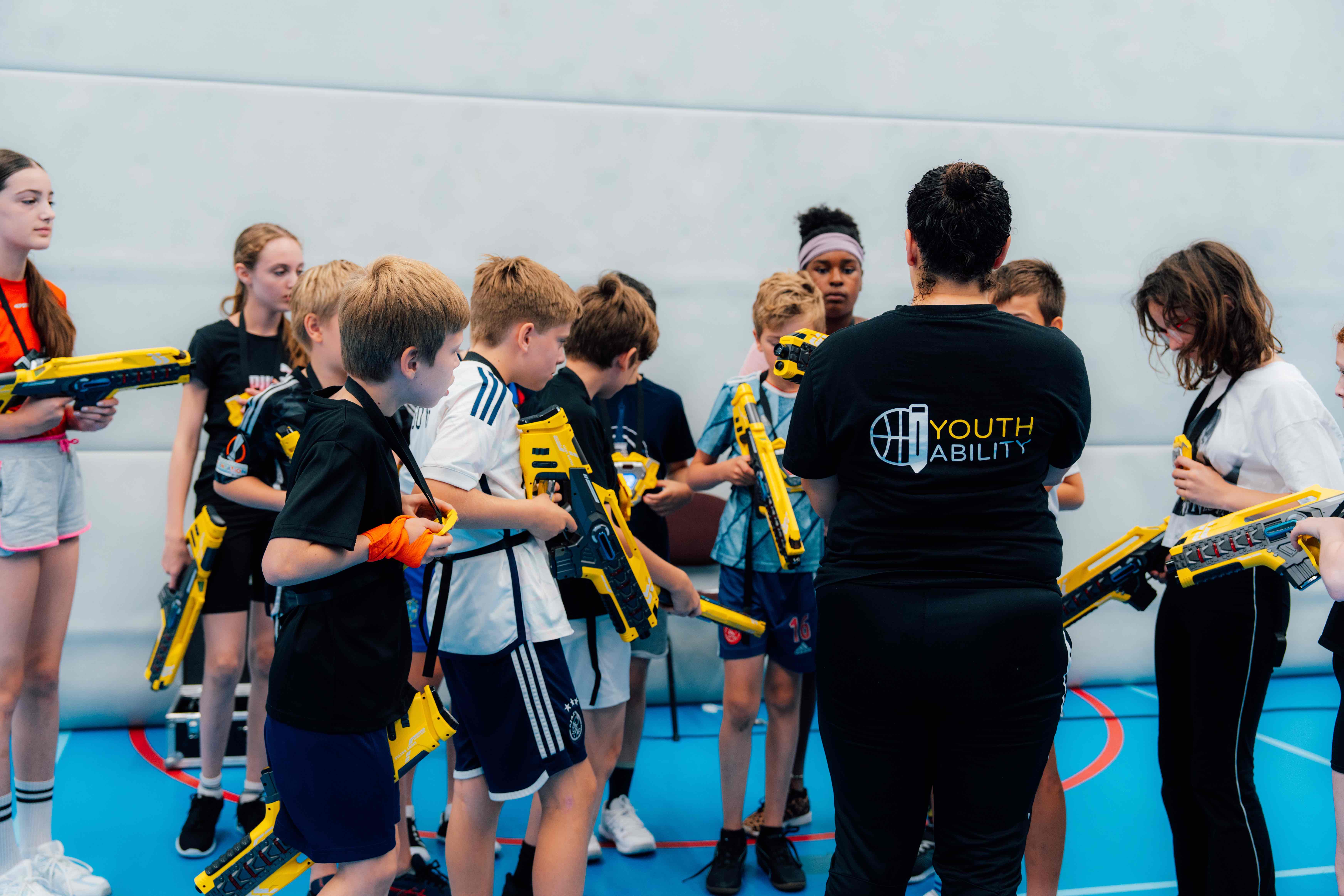 Groep kinderen in sportkleding in een zaal verzamelen rondom een instructeur met een T-shirt met opschrift 'Youth Ability', terwijl ze gele lasergame-wapens vasthouden.