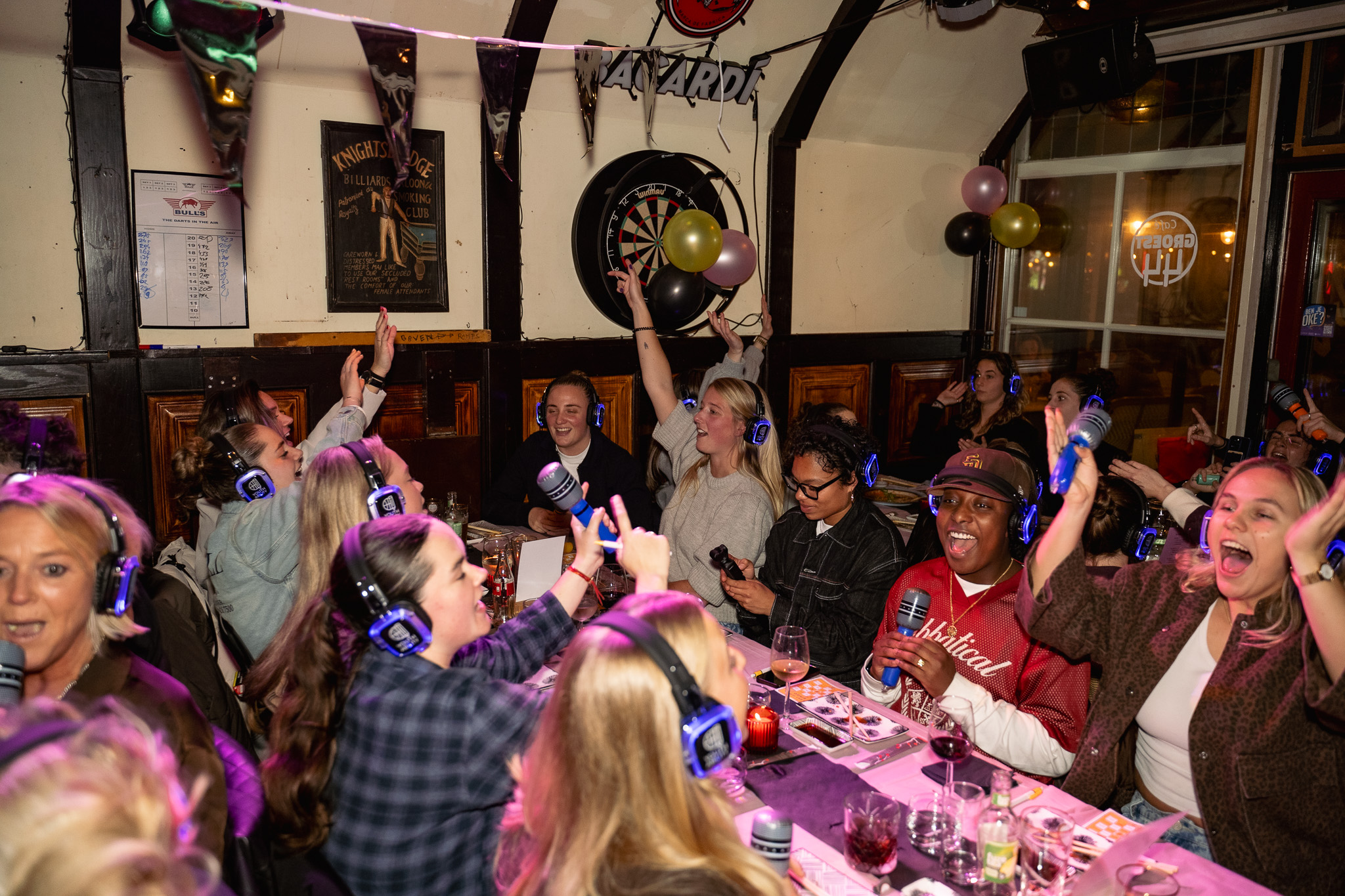 Groep mensen met koptelefoons zingen en vieren tijdens een stille disco in een gezellige kroeg met ballonnen aan de wand.