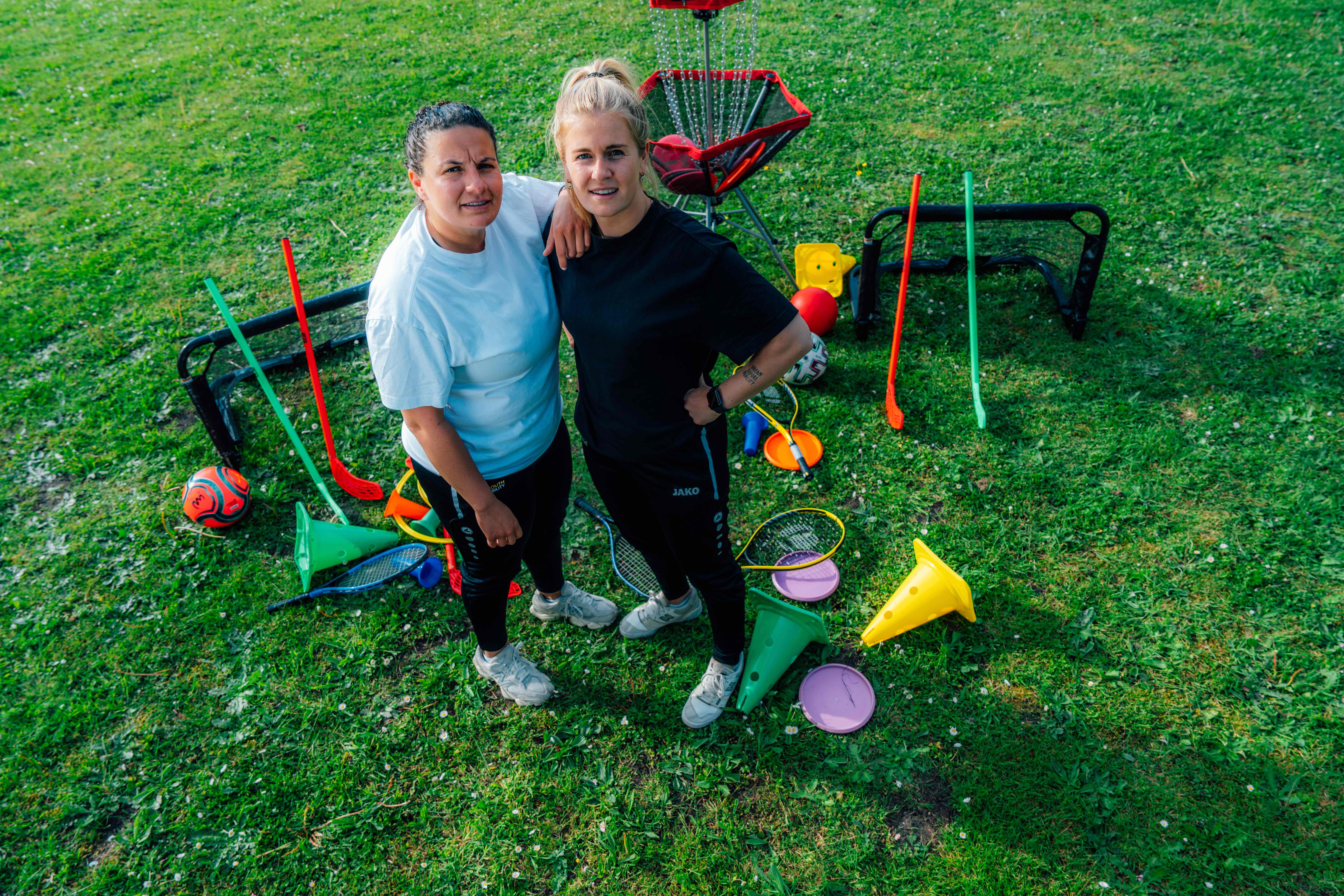 Twee vrouwen op een grasveld omringd door sport- en spelmaterialen, inclusief een voetbal, lacrosse sticks, kegels en een disc golf mand.