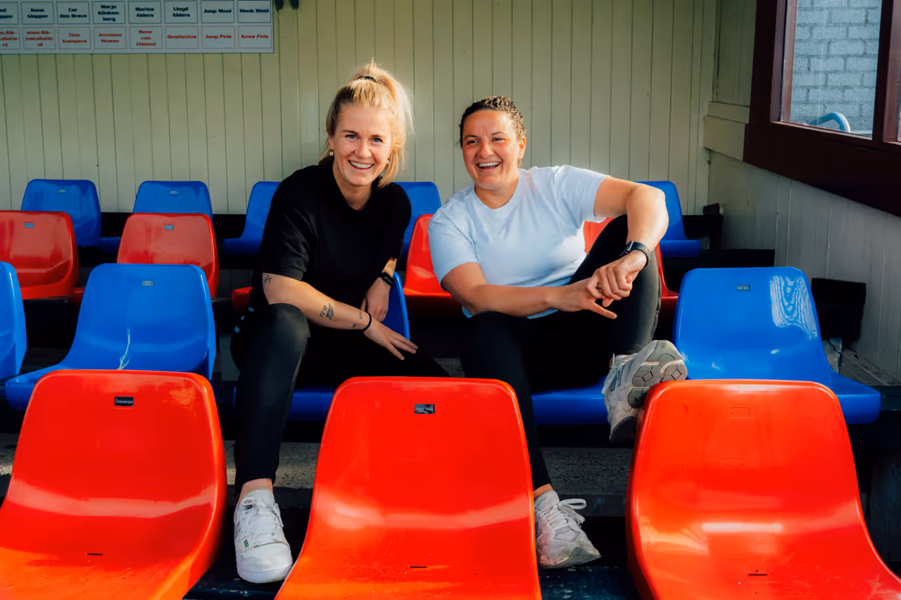 Twee glimlachende vrouwen zittend op rode en blauwe plastic stoelen in een overdekte tribune.