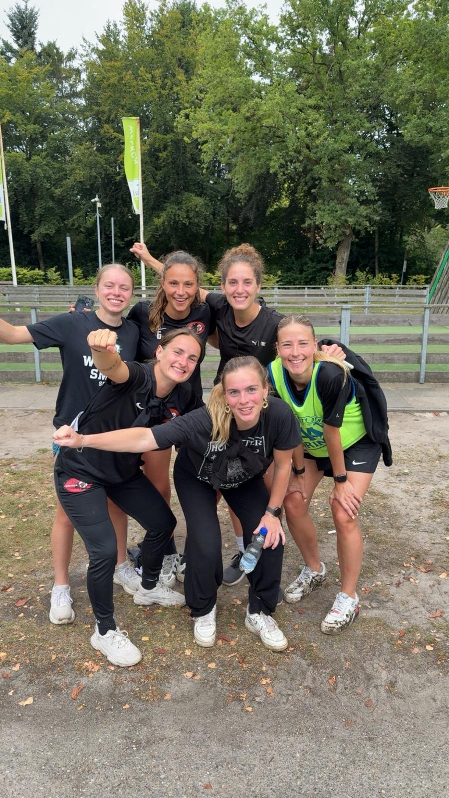 Zes lachende jonge vrouwen in sportkleding poseren buiten op een grasveld met bomen op de achtergrond.