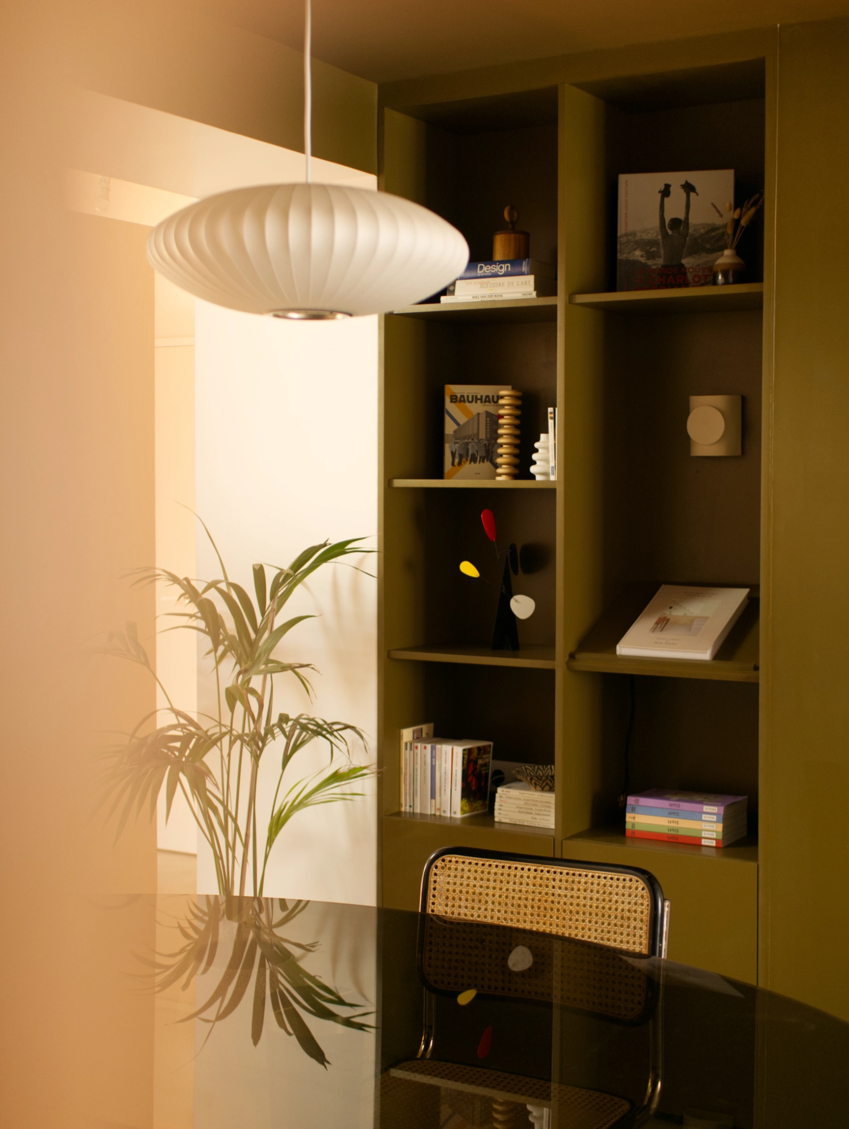 Photo de l'espace repas composé d'une table ovale en verre fumé au premier plan et d'une bibliothèque sur mesure peinte en vert mousse. De jolis objets de décoration habillent les niches ouvertes de la bibliothèque en plus des livres. Au plafond, la suspension bubble saucer de Hay contraste avec le plafond peint dans la même teinte que la bibliothèque en arrière-plan. Le cadrage de la photo permet de réaliser le contraste entre le côté très chaleureux de l'espace repas et le côté très lumineux de la cuisine que l'on devine en arrière-plan.