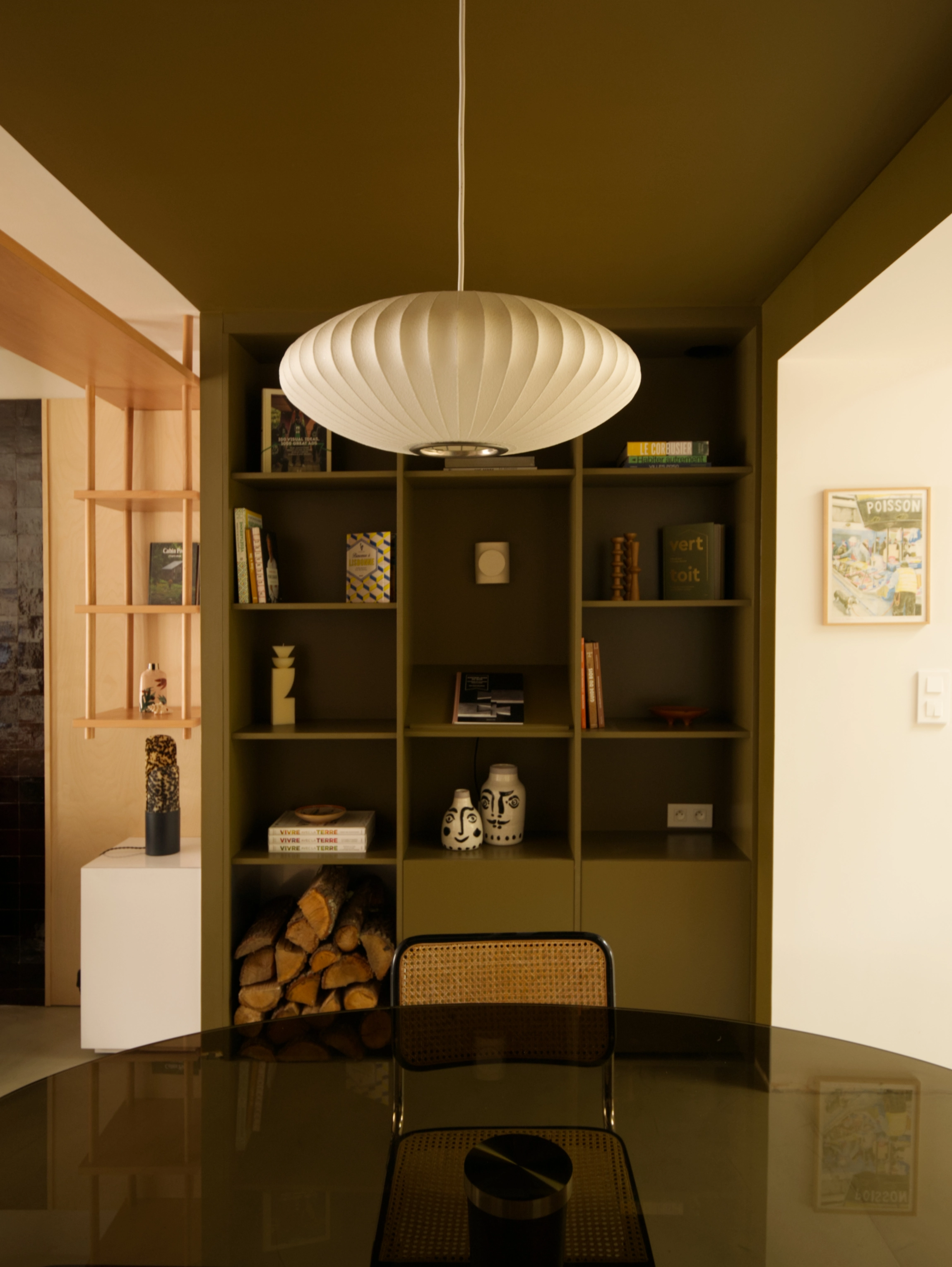 Photo de l'espace repas composé d'une table ovale en verre fumé au premier plan et d'une bibliothèque sur mesure peinte en vert mousse. De jolis objets de décoration habillent les niches ouvertes de la bibliothèque en plus des livres. Au plafond, la suspension bubble saucer de Hay contraste avec le plafond peint dans la même teinte que la bibliothèque en arrière-plan. L'ambiance est chaleureuse et feutrée.