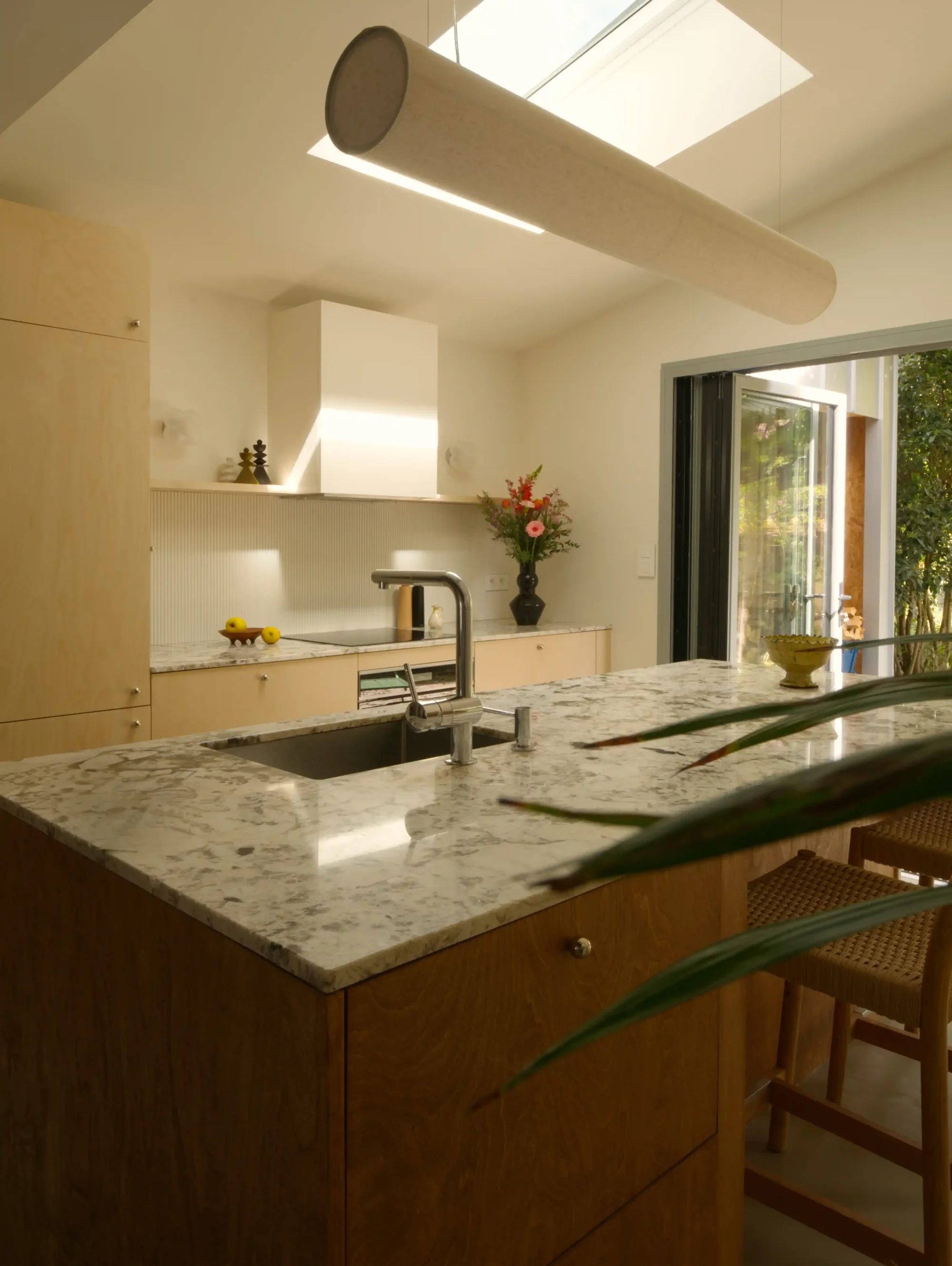 Vue de l'îlot central de la cuisine du projet Leloyer. L'îlot en bois est rehaussé d'un plan de travail en granit dans les tons beige. Avec ses deux essences de bois, la cuisine est très chaleureuse. Un puit de lumière vient rendre l'atmosphère très chaleureuse.