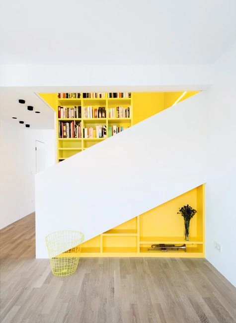L'escalier est caché derrière un garde corps maçonné de couleur blanche. En dessous, l'espace perdu est habillé d'une bibliothèque ouverte peinte en jaune. Au fond, sur le mur de la cage d'escalier, on retrouve la continuité de la bibliothèque jaune.