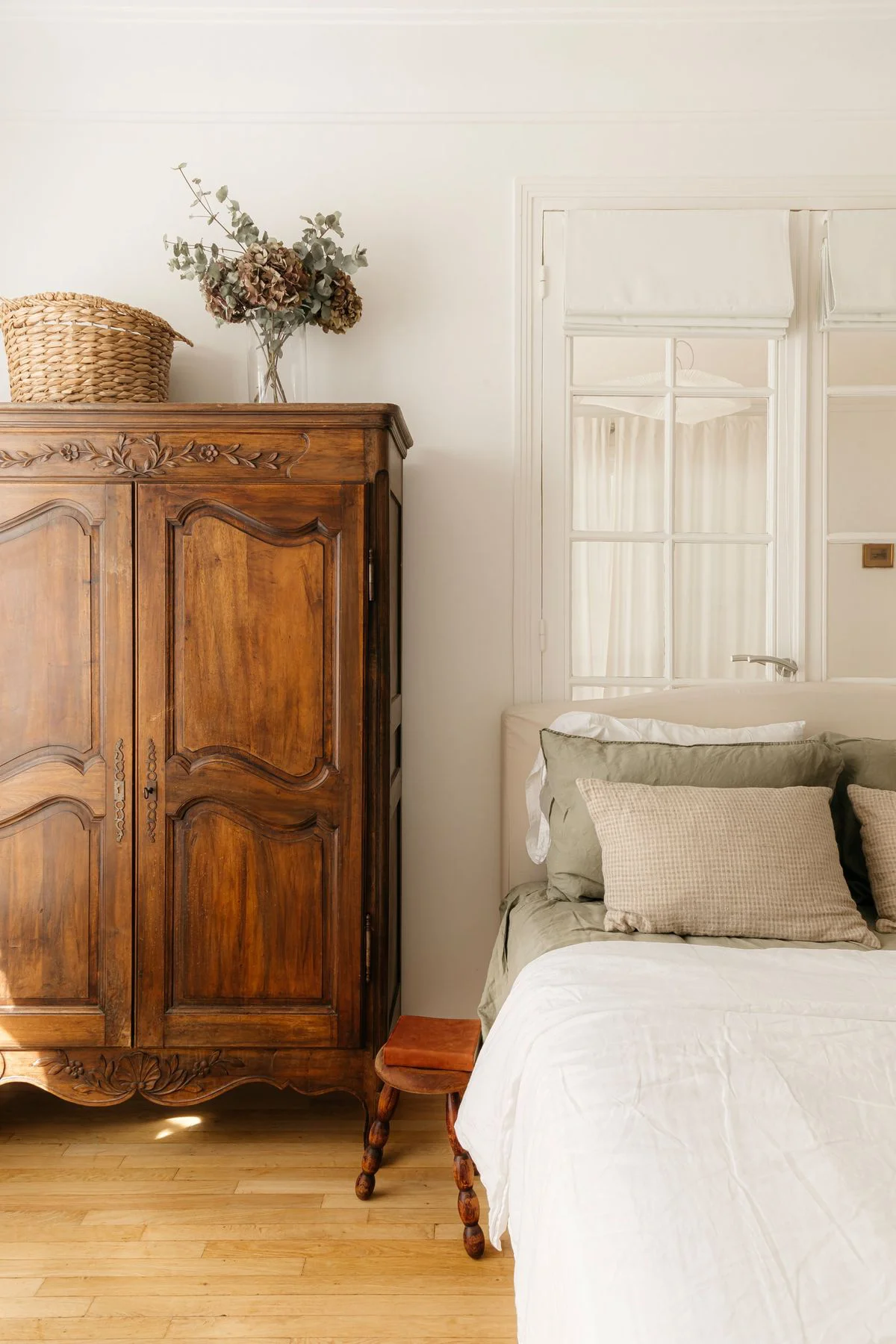 Dans cette chambre, une armoire rustique en bois sculpté se place à côté d’un lit habillé de linge clair et de murs aux teintes neutres.