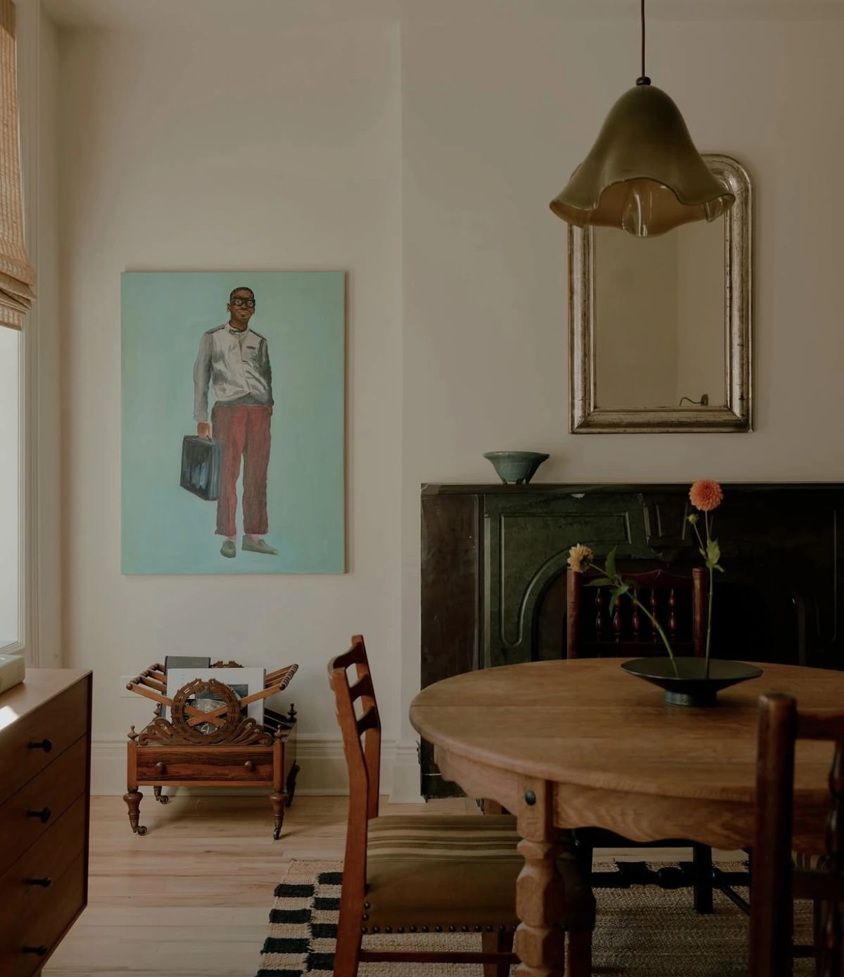 Une table ronde ancienne en bois occupe la salle à manger, accompagnée de chaises vintage, d’un miroir patiné et d’un tableau figuratif très contemporain, accroché au mur.