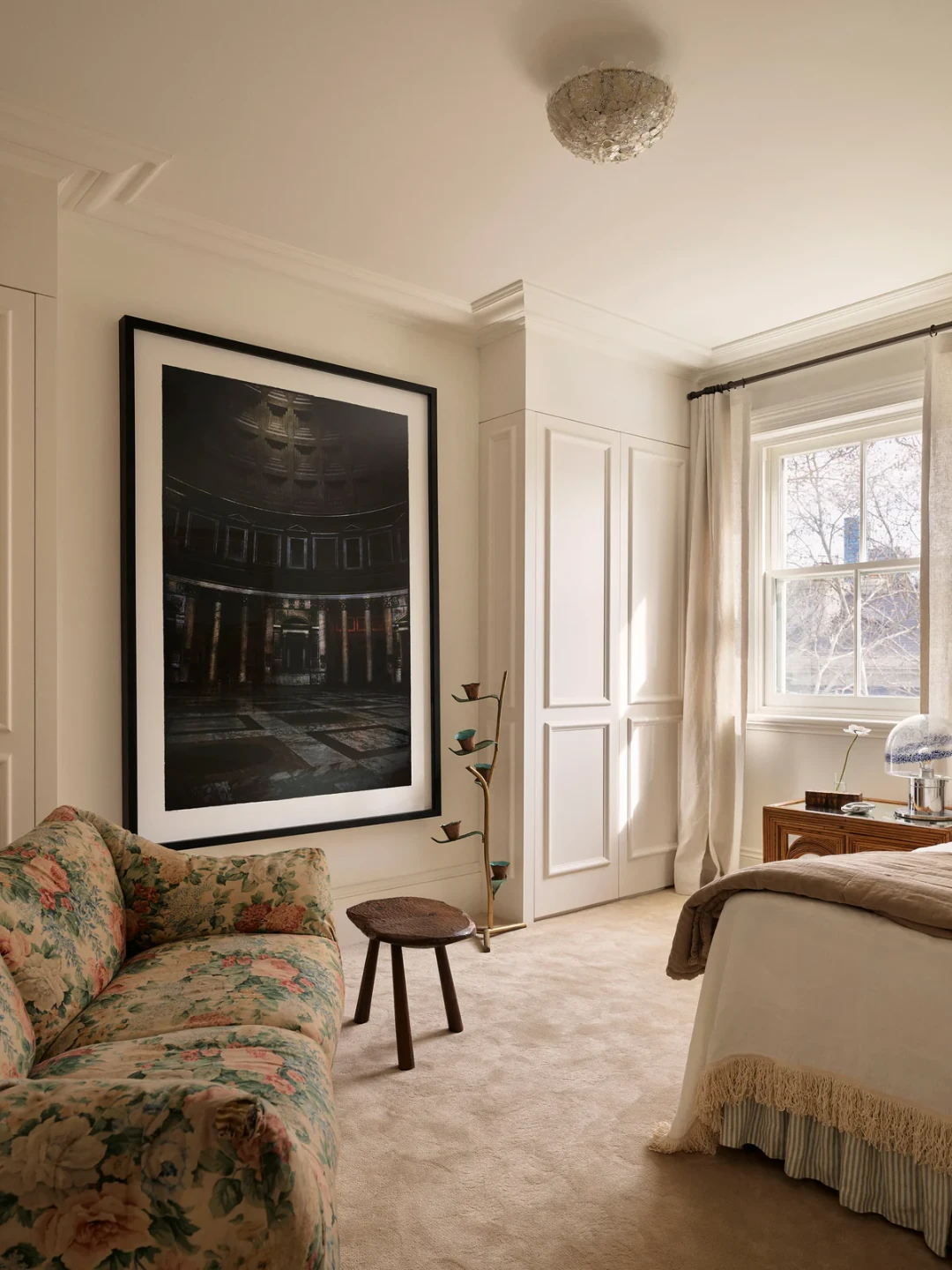 Un canapé désuet aux motifs fleuris prend place dans une chambre claire, sous une très grande photographie contemporaine encadrée et à côté d’un petit tabouret rustique en bois.