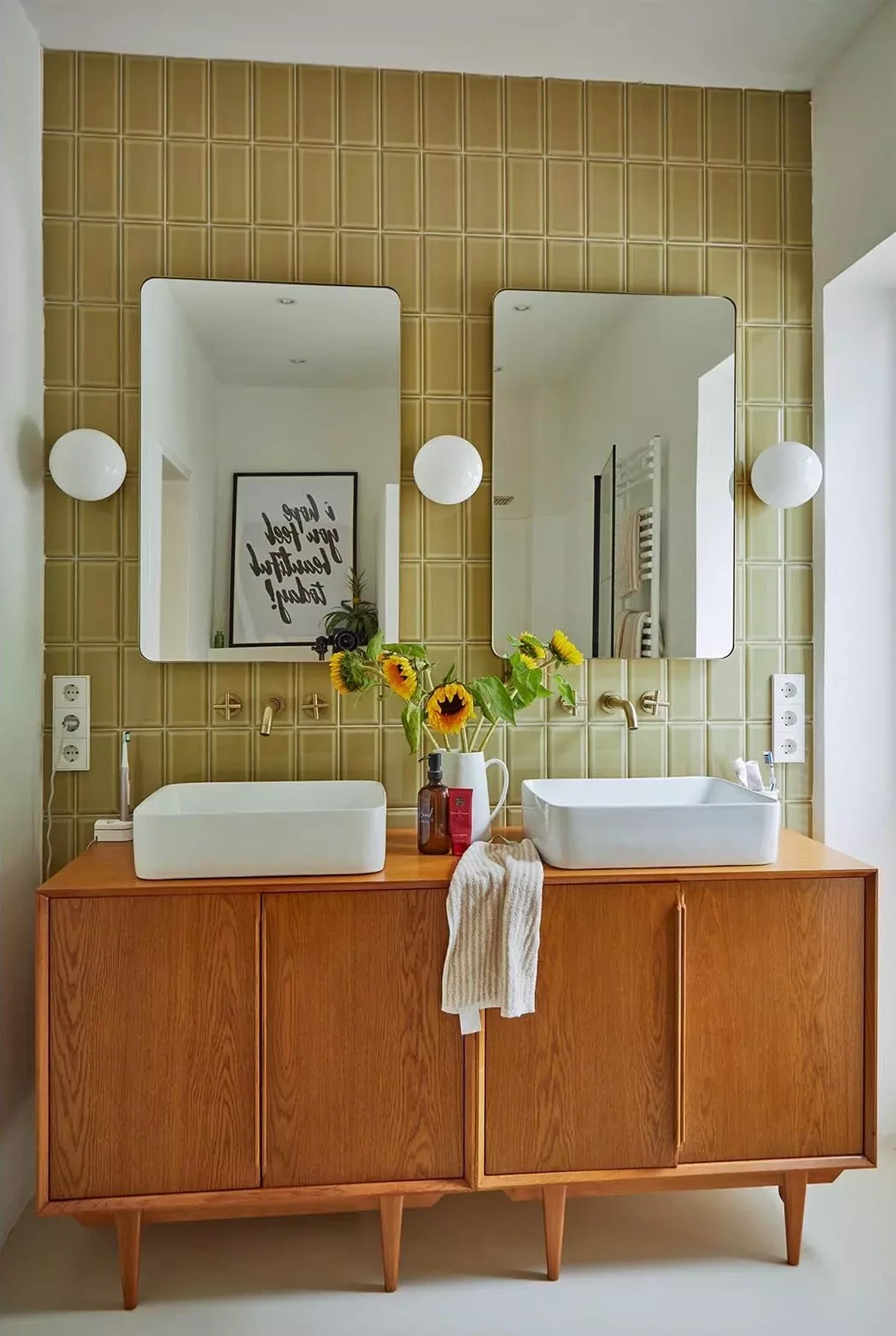 Cette enfilade des années 50 supporte deux vasques en céramique devant un mur carrelé coloré et des miroirs épurés, associant matériaux nobles et design contemporain.