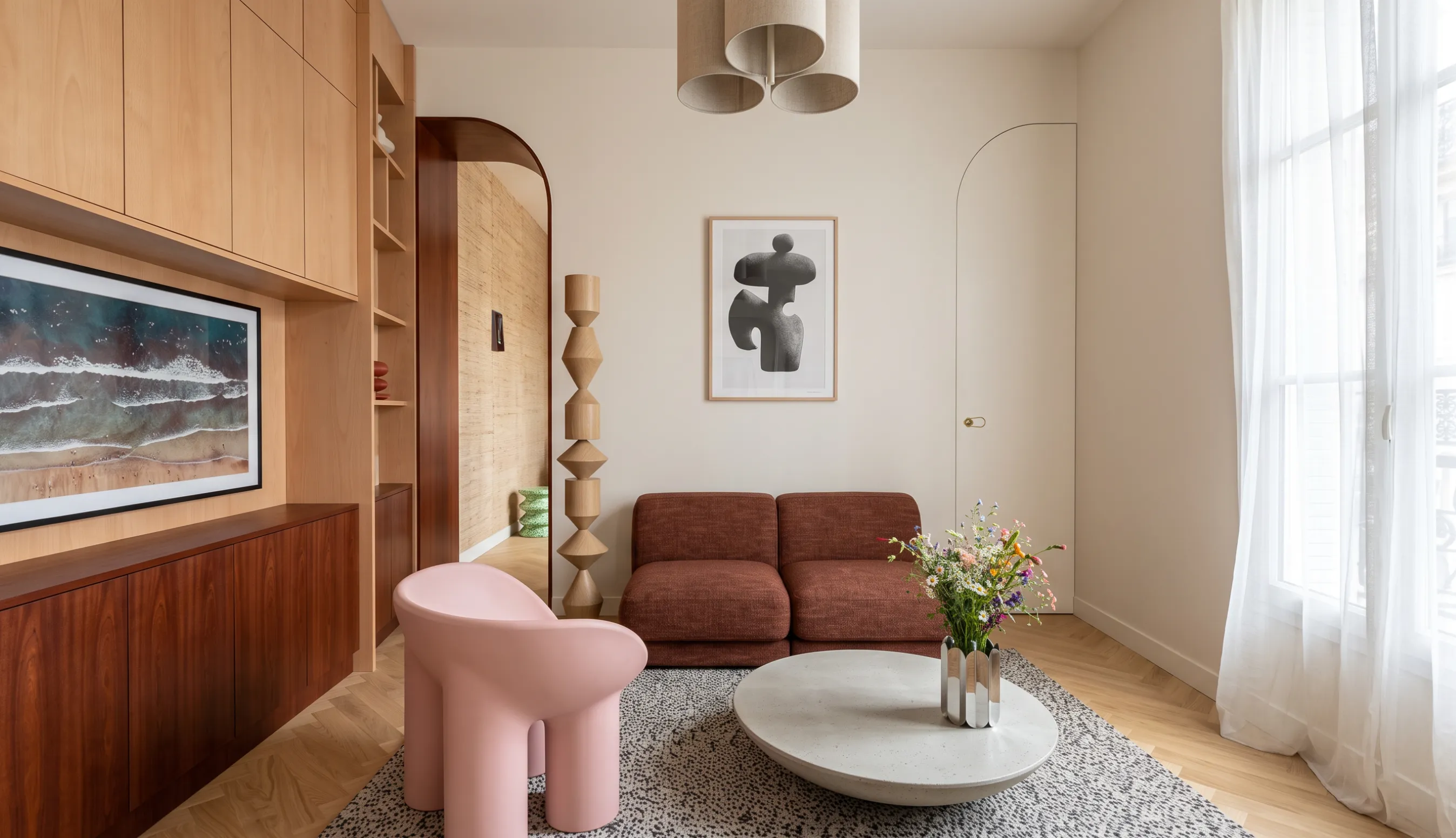 Salon en camaïeu terracotta : canapé modulable bordeaux, fauteuil rose, table basse ronde en pierre claire, bibliothèque sur mesure en bois clair et arche en acajou ouvrant sur un couloir habillé de raphia.