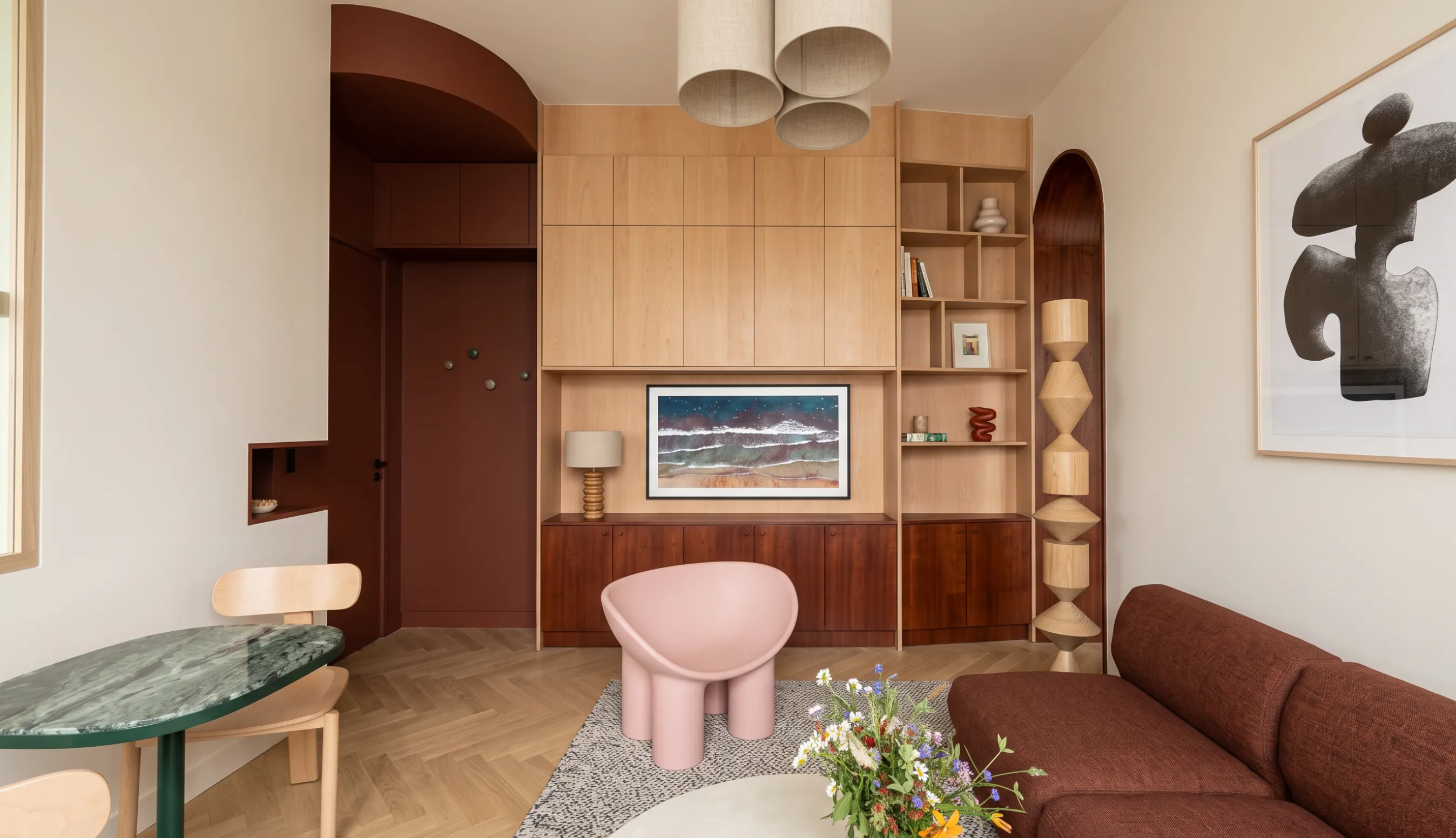 Meuble bibliothèque sur mesure en bois clair et acajou intégrant écran, rangements fermés et niches ouvertes, face à une table de repas en marbre vert et à l'arche bordeaux de l'entrée.
