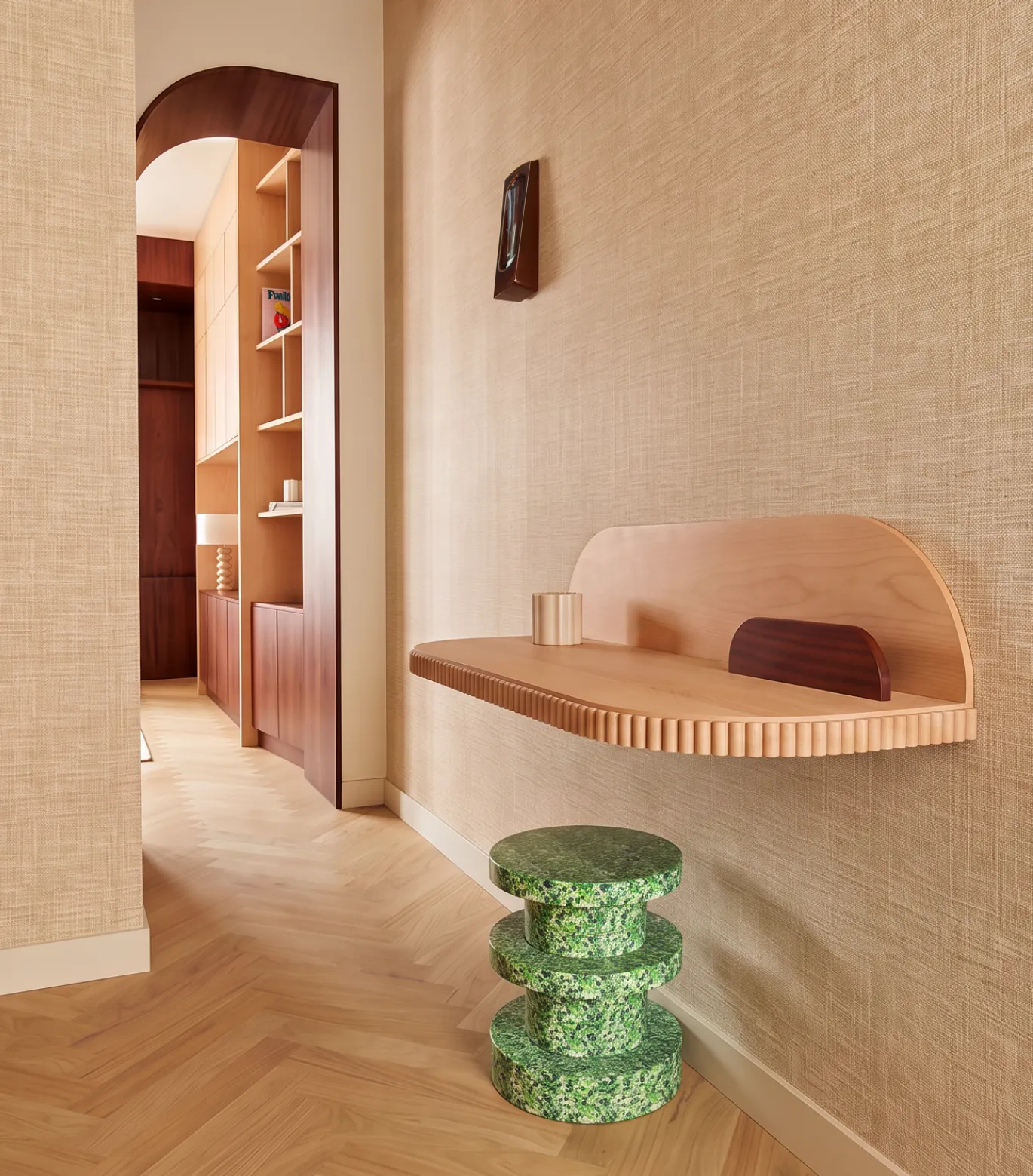 Bureau mural sur mesure en bois clair à façade cannelée, fixé sur un mur tendu de raphia, avec tabouret en pierre verte mouchetée et parquet chevron.