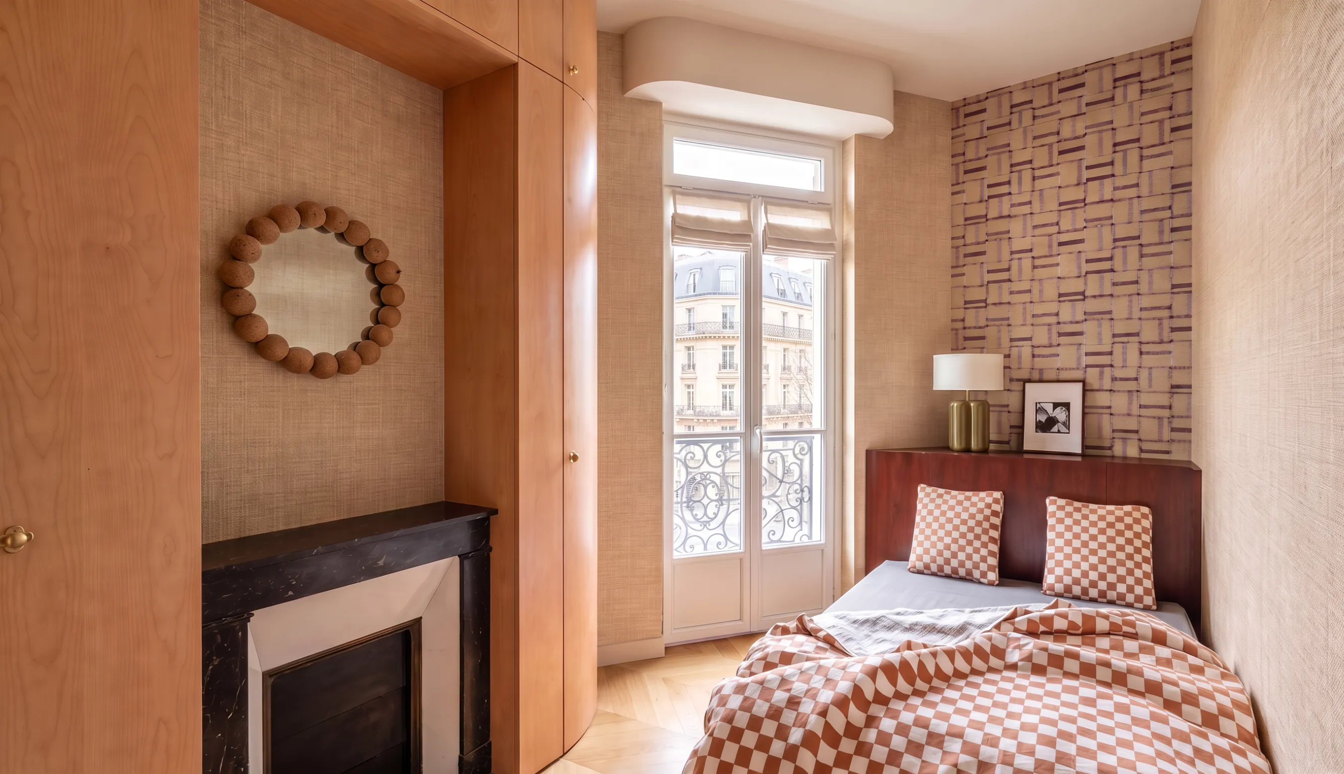 Chambre d'appartement haussmannien : cheminée en marbre noir, miroir rond en terre cuite, placards courbes en bois clair, papier peint à motif tressé et tête de lit en acajou avec linge de lit à damier terracotta.