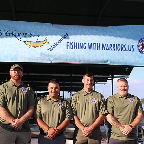 Four veterans in uniform standing under Fishing with Warriors welcome banner