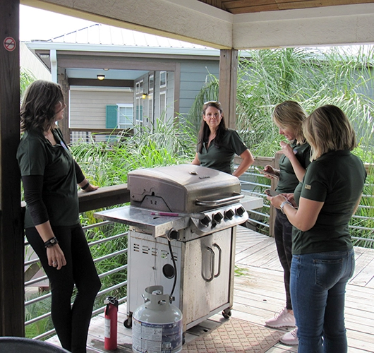 Four women in uniform gather around a grill on an outdoor deck