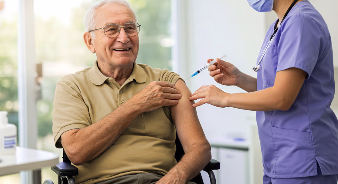 Smiling elderly man in a wheelchair receiving a vaccination from a healthcare worker in purple scrubs.