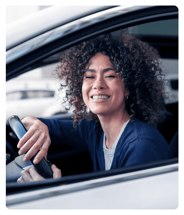Mulher negra, de cabelos cacheados, sorrindo, com blusa azul. Está olhando para frente, com as mãos no volante do carro.