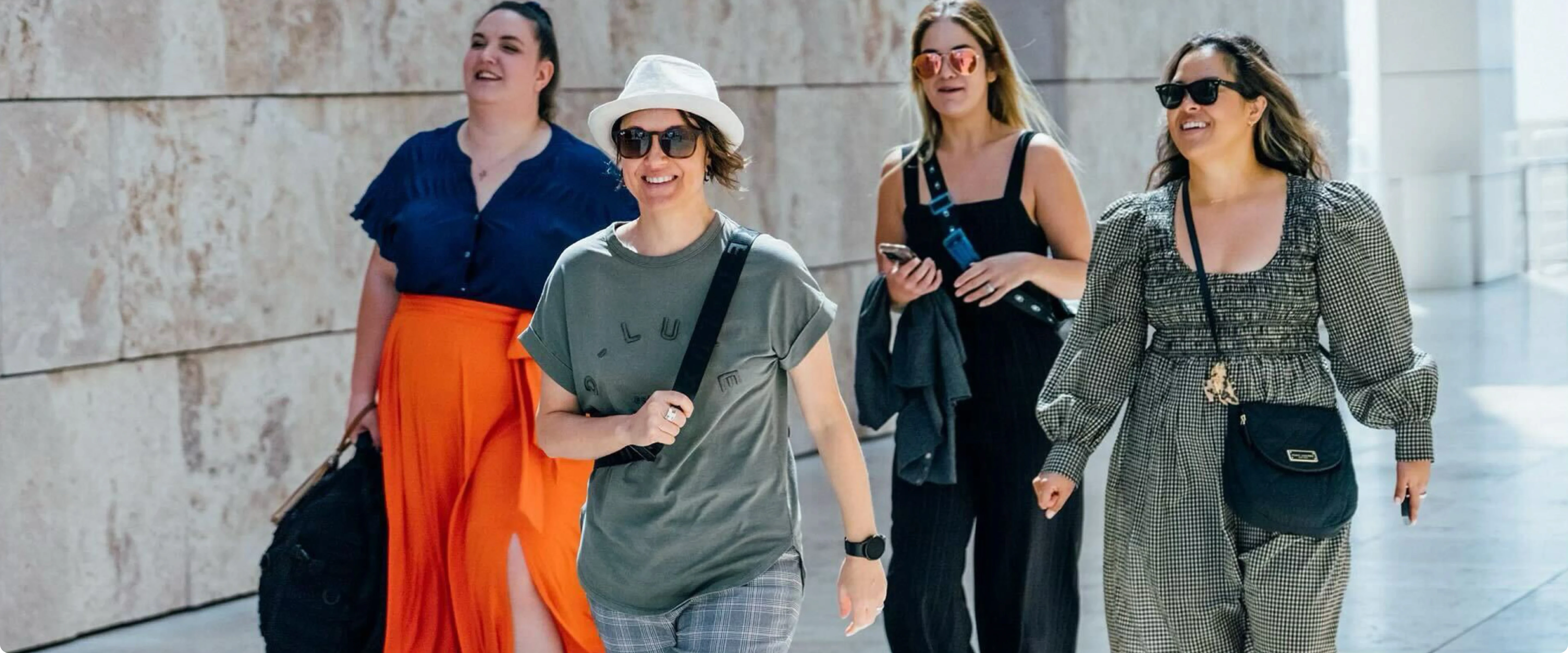 A group of people walking outdoors on a sunny day, smiling and chatting as they pass a stone wall, suggesting camaraderie and inclusivity.