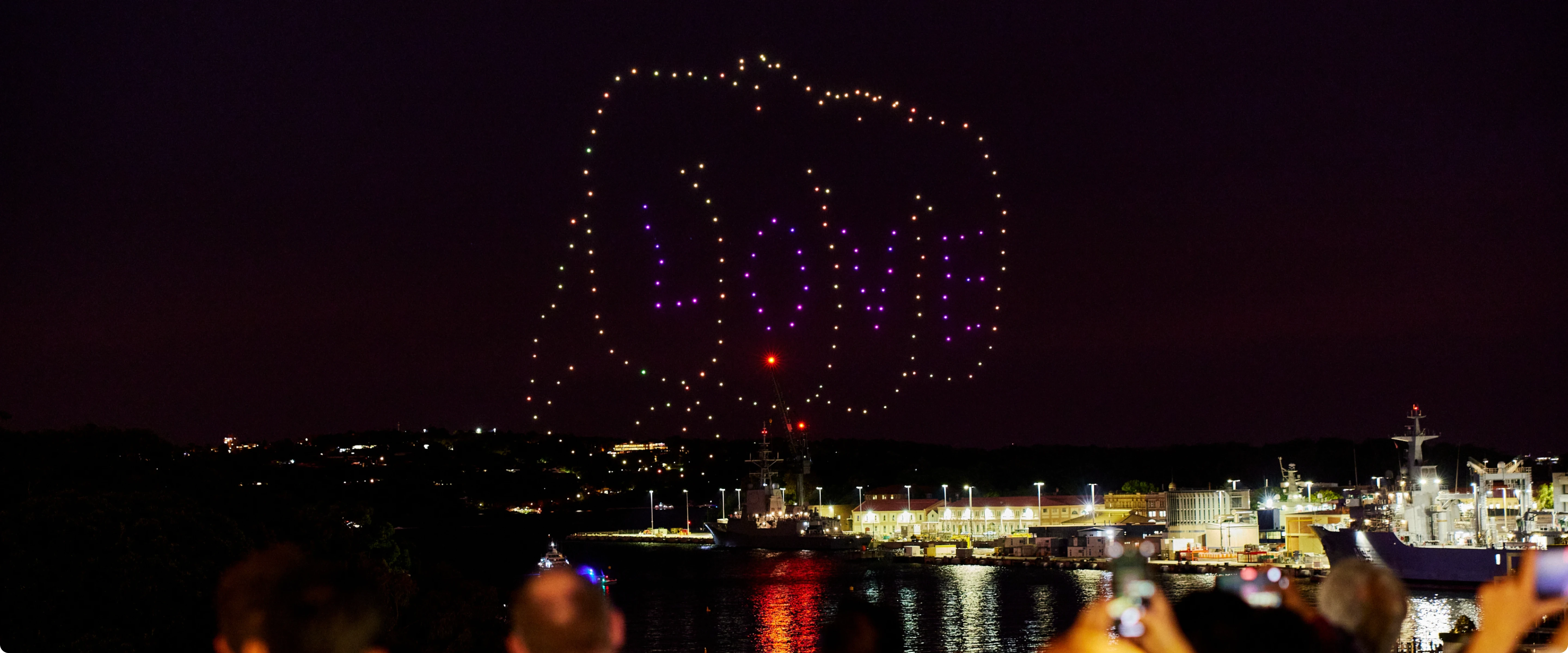 Crowd watching a night-time drone light show spelling out the word “LOVE” in the sky.