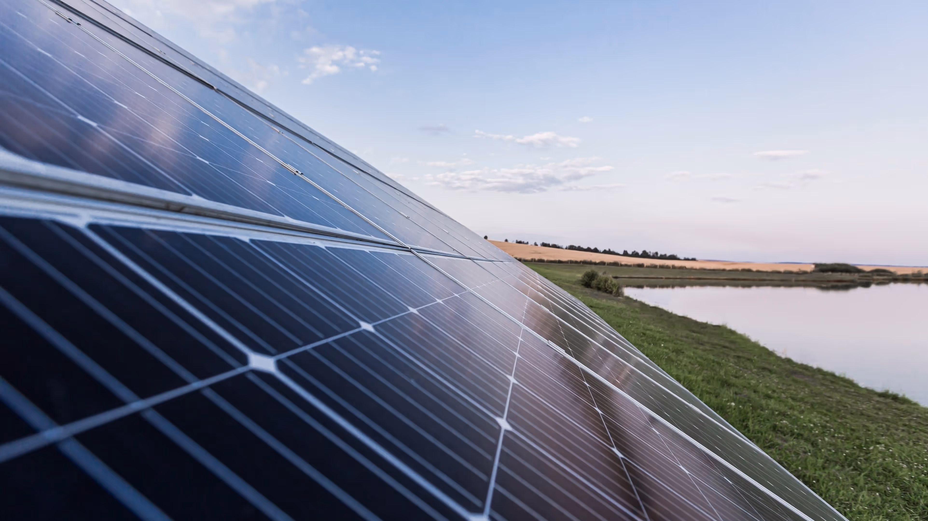 Close-up of solar panels angled towards the sky with a grassy field and pond in the background.