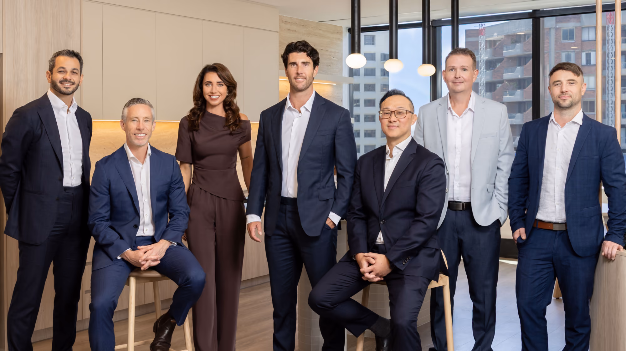 Group of seven professionally dressed people posing in a modern office with large windows and wooden accents; four men standing and one sitting on a stool on left, one woman standing in the center, one man sitting on a stool on the right.