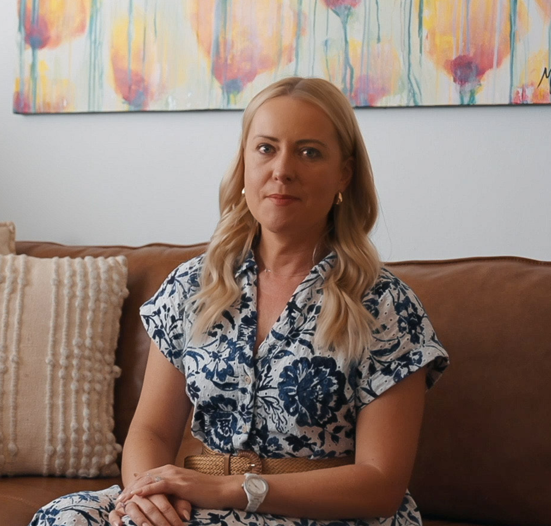 Blonde woman with wavy hair sitting on a brown couch wearing a white and blue floral dress, with a beige belt and a white watch, a textured beige pillow and colorful abstract painting in the background.