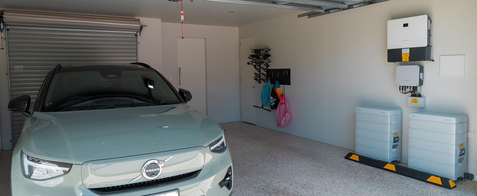 Light green Volvo electric car parked inside a clean, modern garage with skateboards on a wall rack and white energy storage batteries installed.