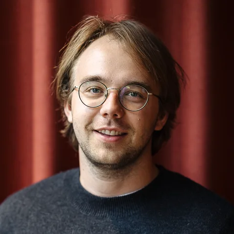 Ung mann med brunt hår og briller smiler mot kameraet, foran en rød gardin.