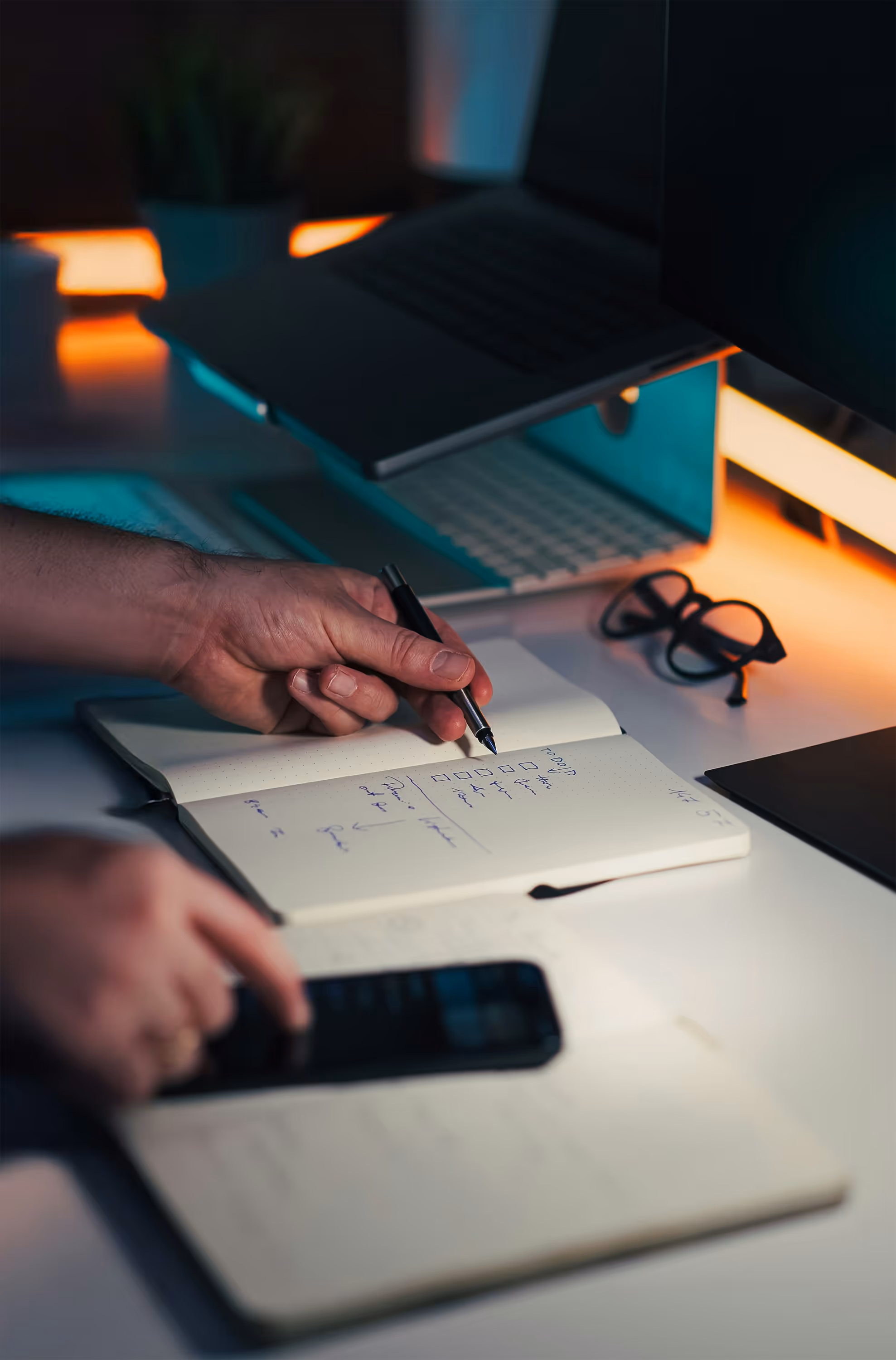 Person écrivant dans un carnet avec un ordinateur portable surélevé, des lunettes et un téléphone sur un bureau éclairé.