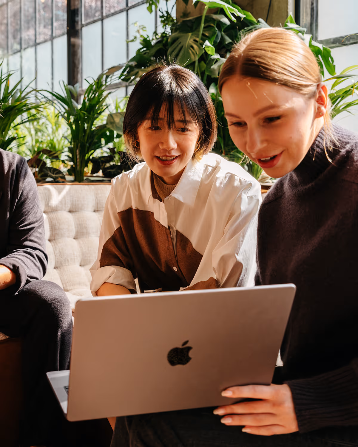 Deux femmes assises sur un canapé regardant un ordinateur portable Apple dans un environnement lumineux avec des plantes en arrière-plan.