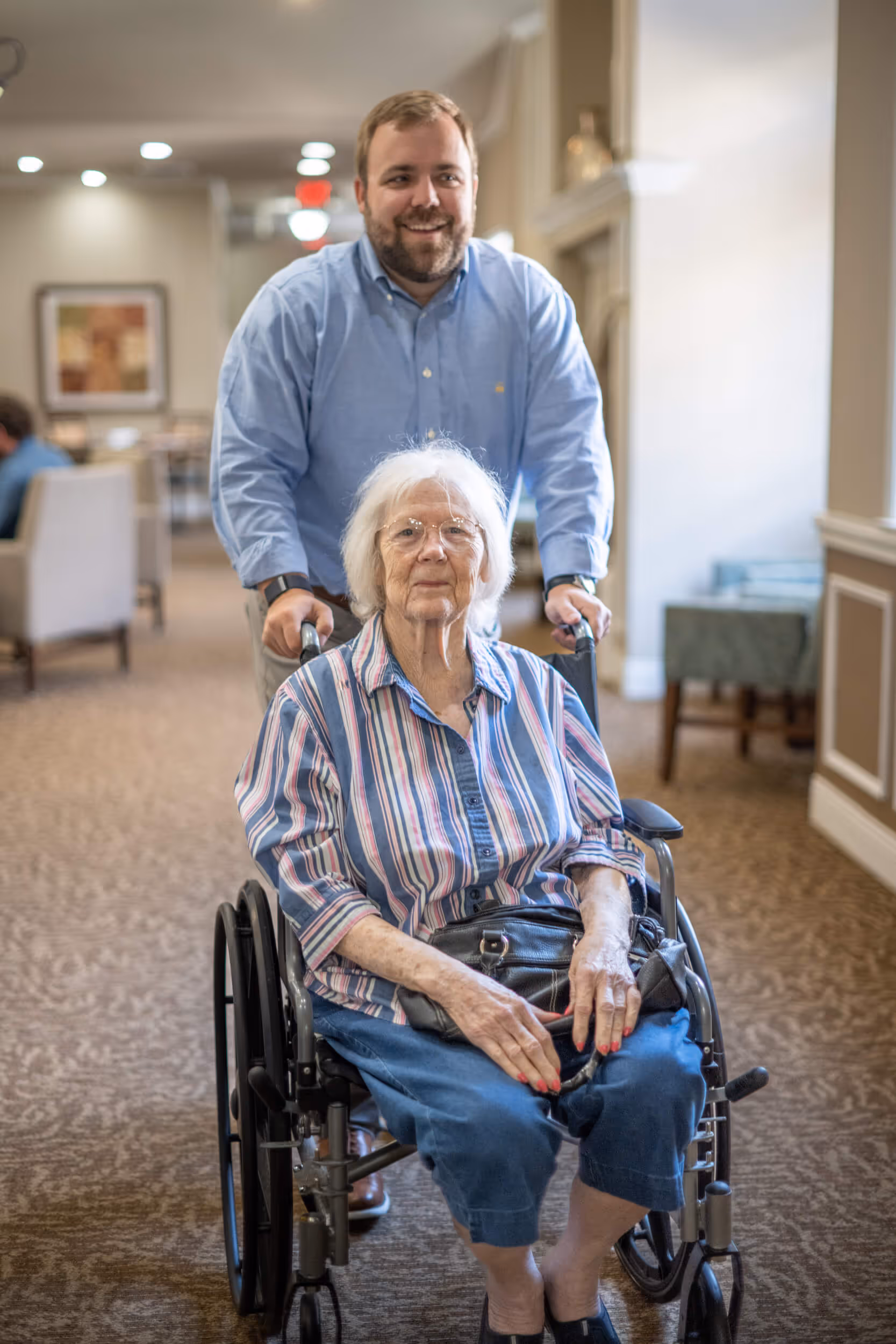 A smiling man in a light blue shirt pushing a senior woman in a wheelchair through a well-lit, cozy facility