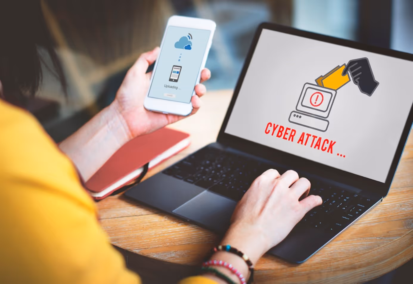 Person hält Smartphone mit Upload-Symbol und nutzt Laptop, auf dessen Bildschirm eine Grafik mit dem Text ‚Cyber Attack‘ und einer Hand, die eine Akte in einen Computer steckt, angezeigt wird.