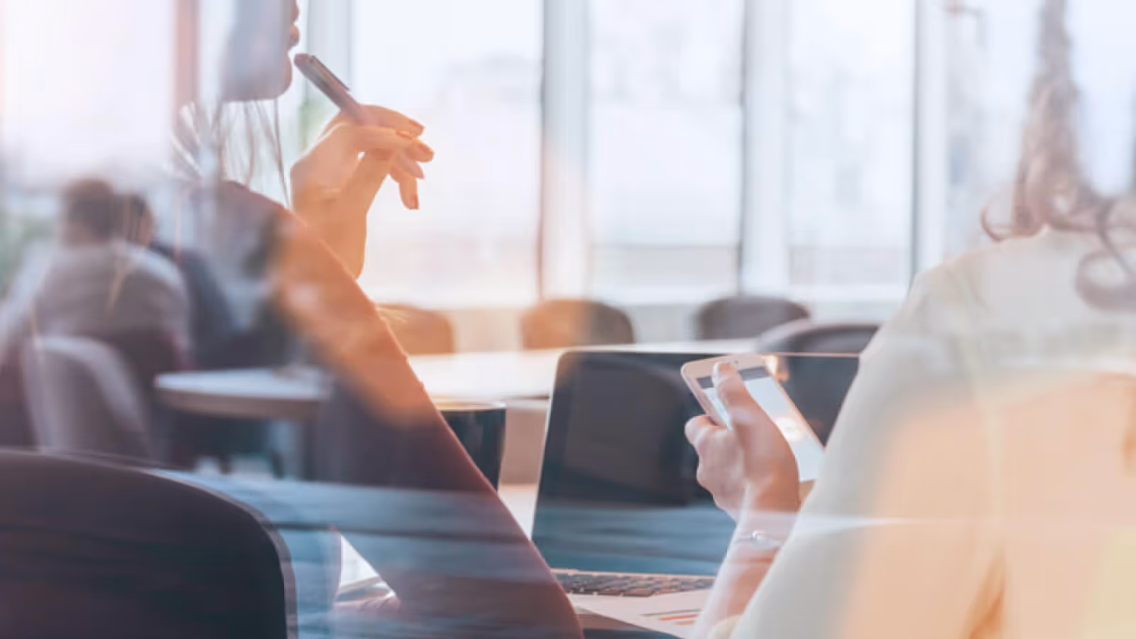 Zwei Personen in einem hellen Büro, eine hält ein Smartphone, die andere einen Stift, mit Laptop und Kaffeetasse auf dem Tisch.