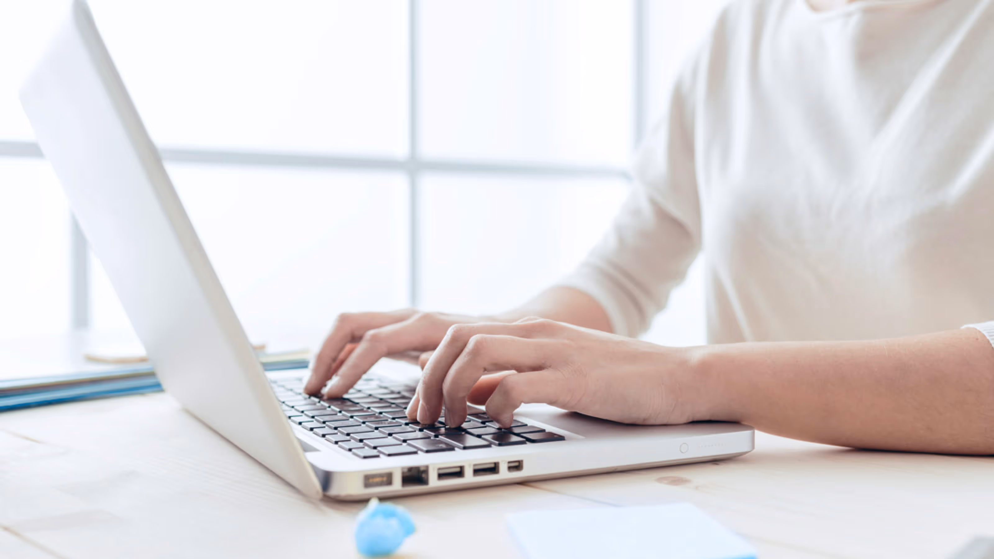 Person tippt auf der Tastatur eines Laptops auf einem weißen Tisch vor einem hellen Fenster.