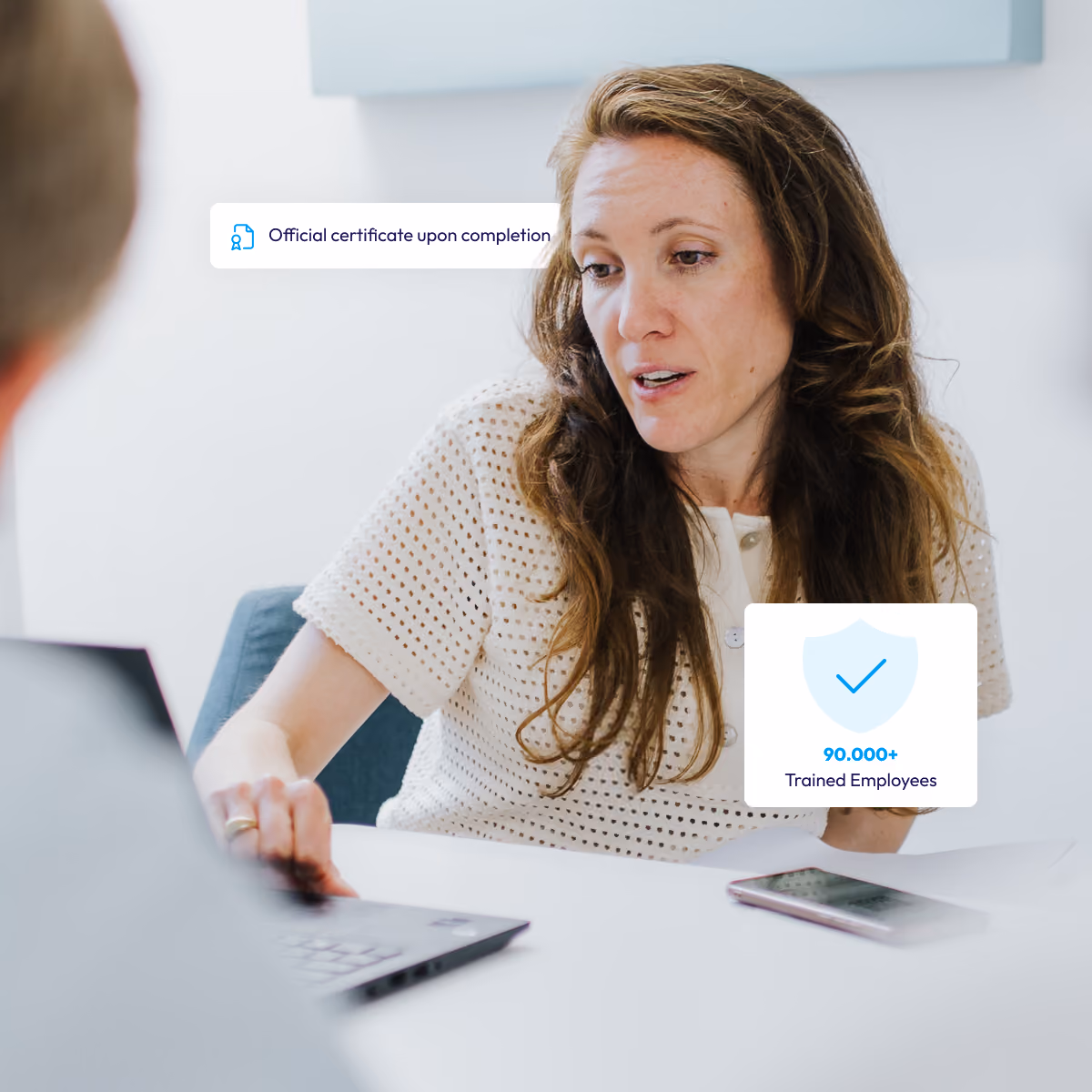 Woman explains something at the table, in front of it laptop and cell phone, text about the certificate and 90,000+ trained employees.