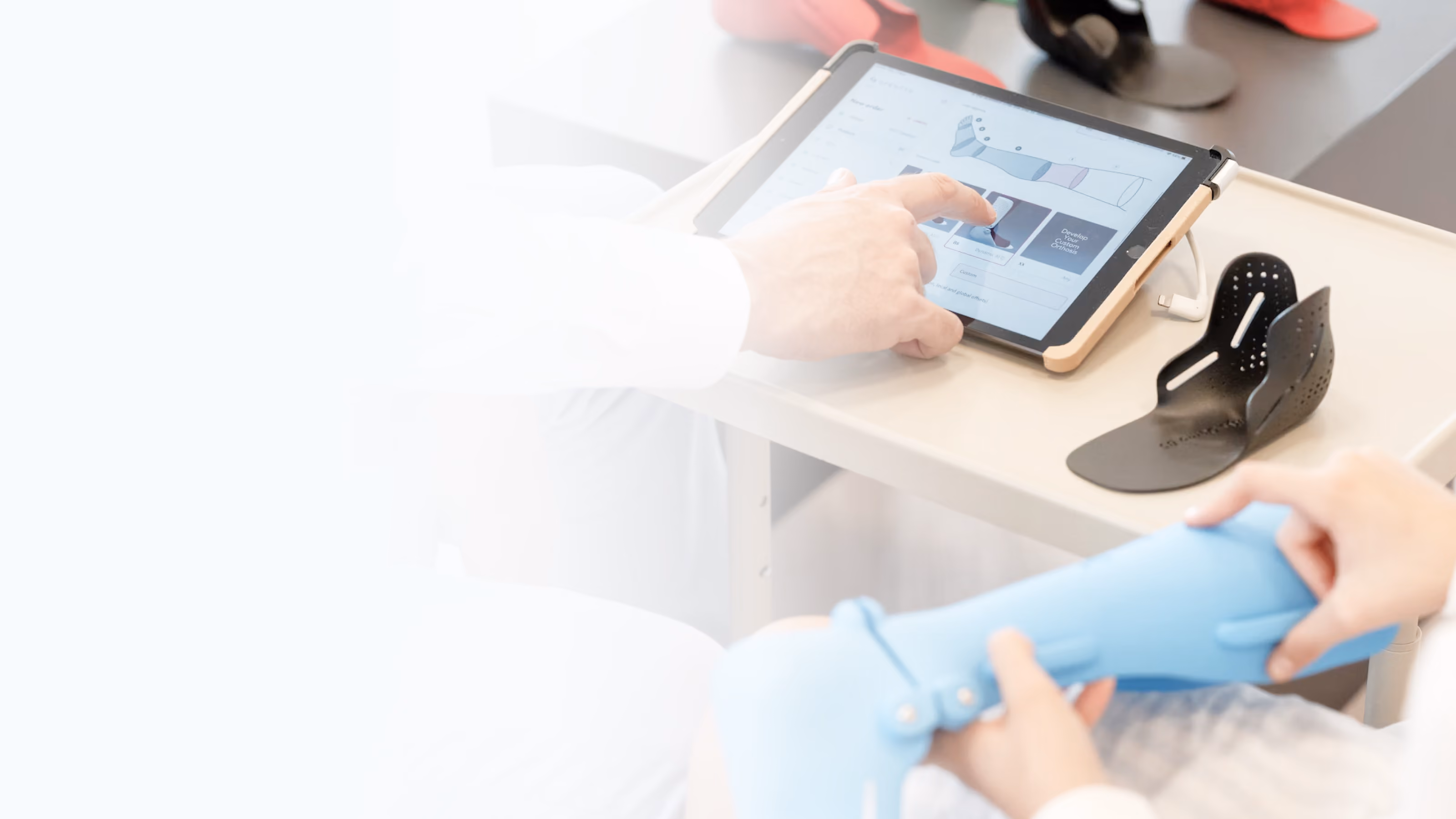 Zwei Personen arbeiten an orthopädischen Hilfsmitteln; eine hält eine blaue Schienenorthese, die andere zeigt auf ein Tablet mit einem technischen Diagramm, daneben liegt eine schwarze Orthese auf dem Tisch.