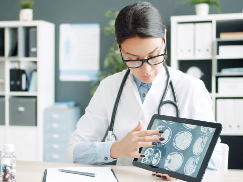 Ärztin in einem weißen Kittel mit Stethoskop um den Hals betrachtet digitale Gehirn-CT-Bilder auf einem Tablet in einer modernen medizinischen Praxis.