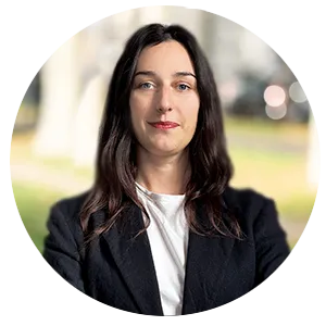 Portrait d'une femme aux cheveux longs et bruns, portant un manteau noir et un t-shirt blanc, avec un fond extérieur flou.
