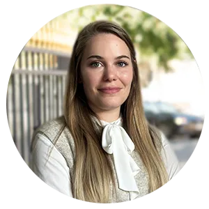 Portrait d'une femme aux cheveux longs blonds, portant une chemise blanche avec un nœud et un gilet beige, en extérieur avec un fond flou d'arbres et de structures urbaines.