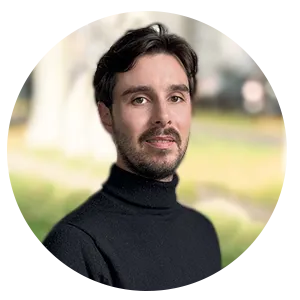 Portrait d'un homme souriant portant un pull noir à col roulé avec un arrière-plan flou extérieur.