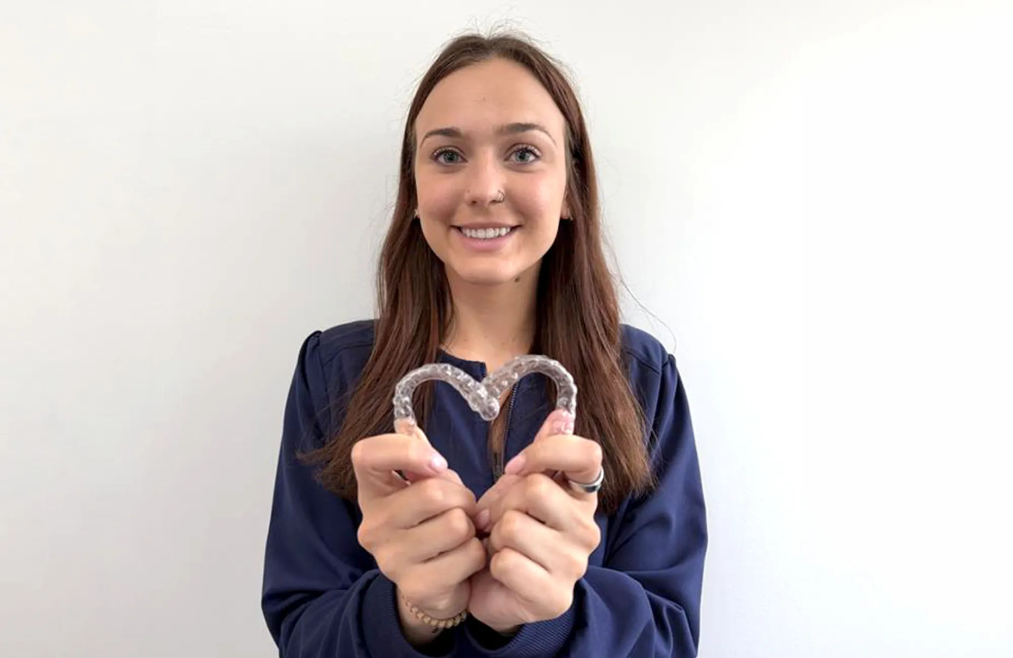 Smiling woman in blue uniform holding a clear dental aligner shaped like a heart.