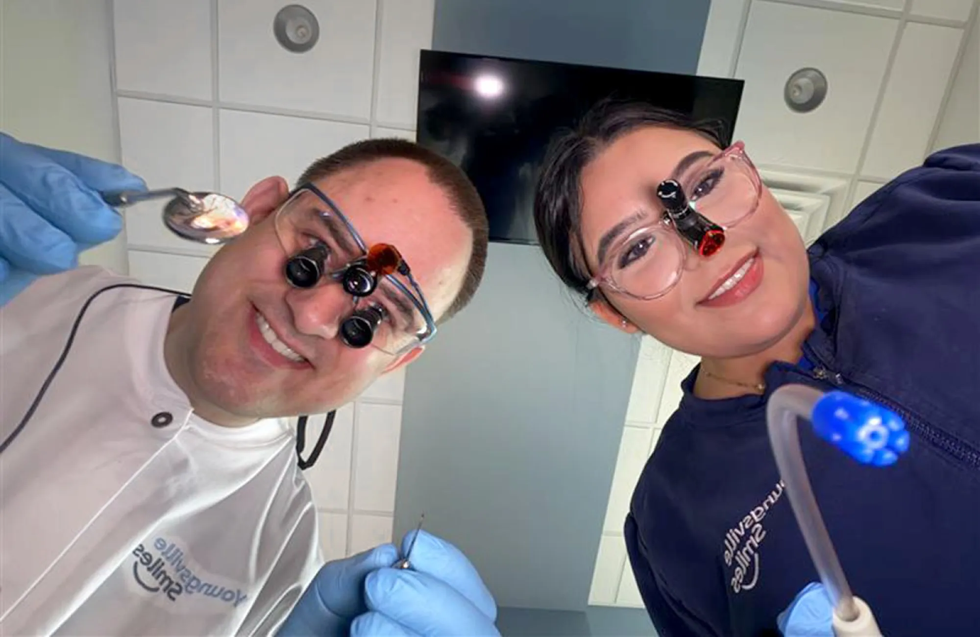 Two smiling dentists wearing magnifying glasses and gloves looking down toward the camera.