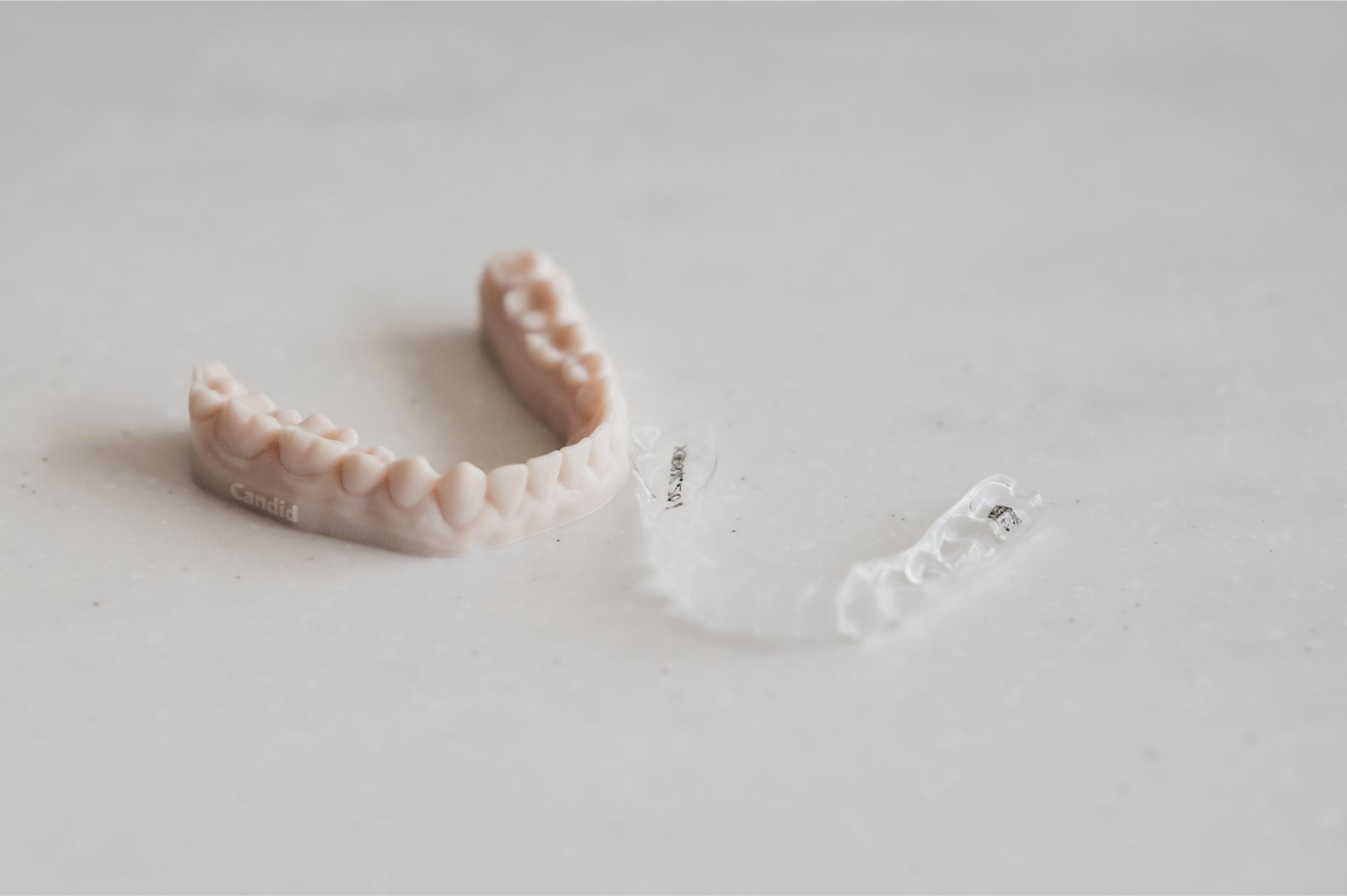 A close-up view of a clear dental aligner next to a beige dental model labeled "Candid," placed on a light-colored surface. The setup highlights orthodontic treatment tools.