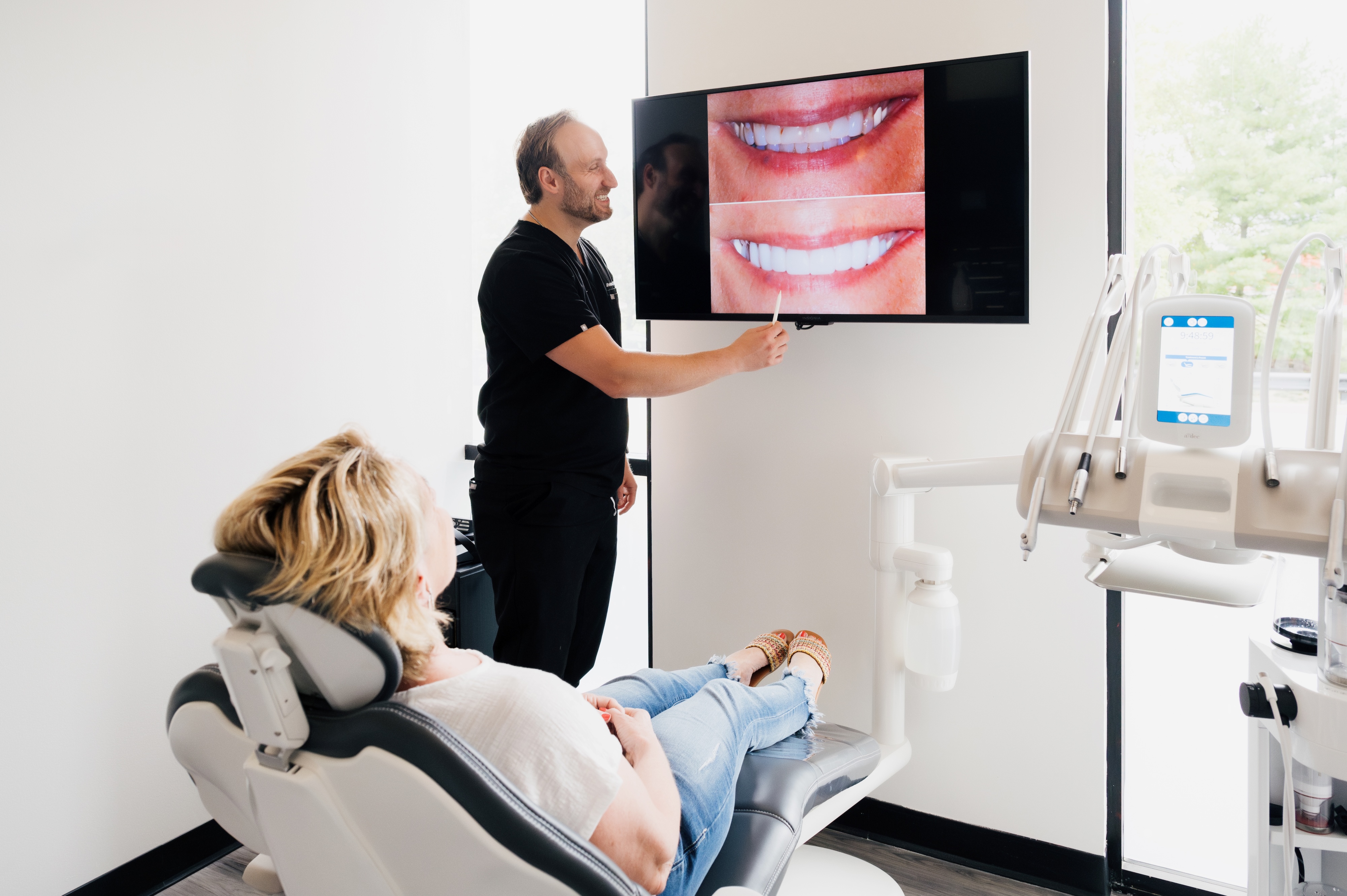 A dental patient is having a final visit for getting veneers and the dentist is showing her on the tv screen her photos of her teeth from before and after getting veneers.
