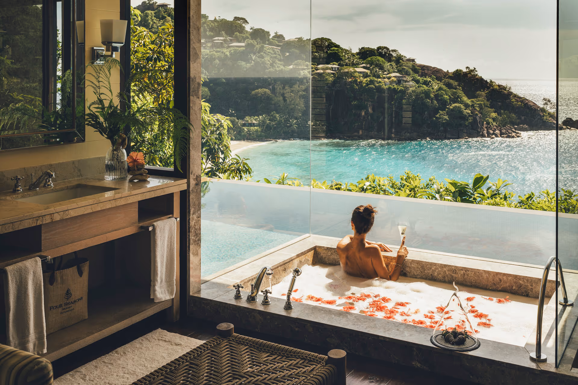 Person relaxing in a bubble bath with flower petals in a modern bathroom overlooking a turquoise sea and lush green hills through large glass windows.
