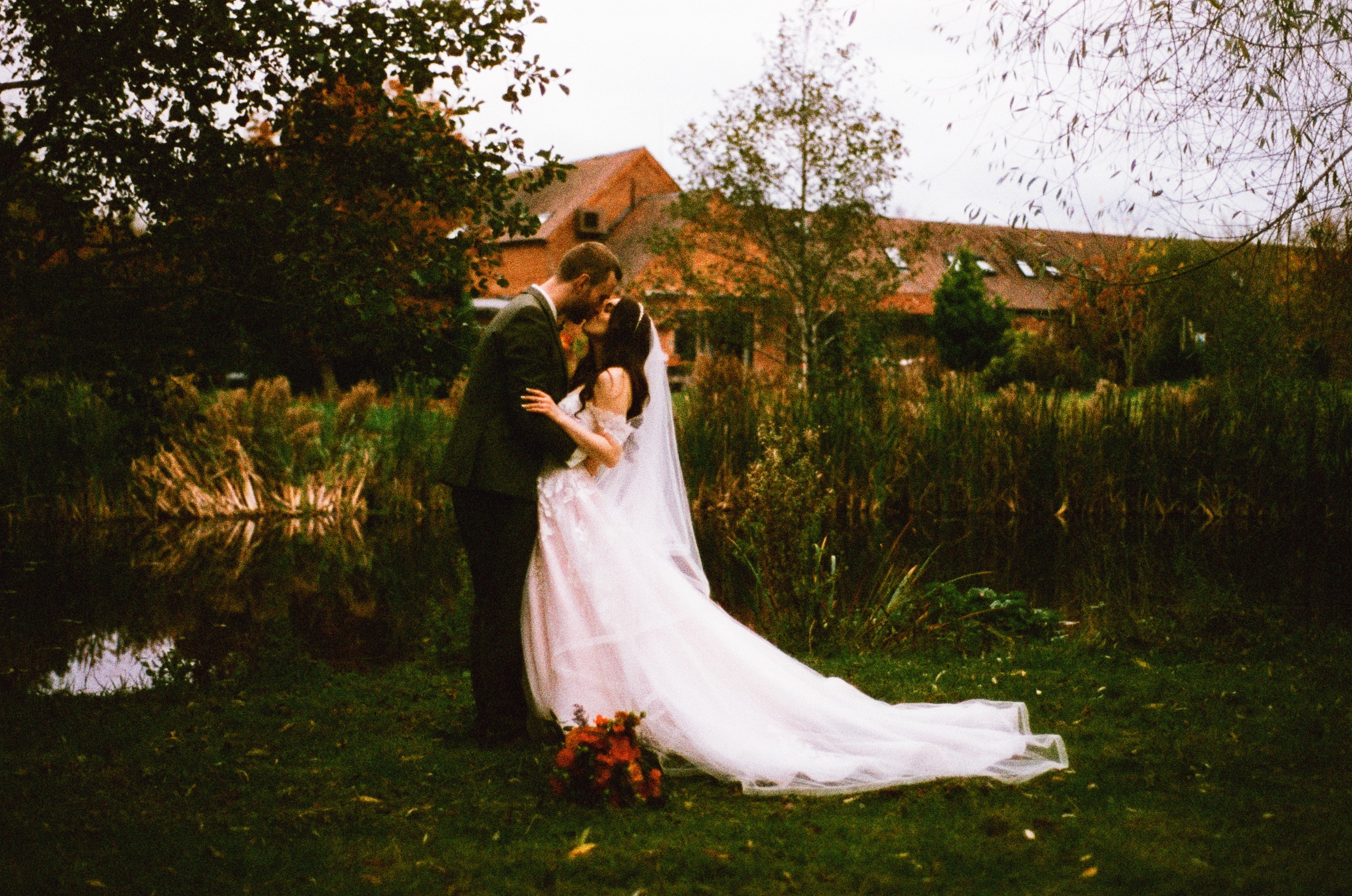 A romantic outdoor wedding scene with a couple kissing near a pond. The bride is wearing a flowing white gown with a long veil, while the groom is in a dark green suit. The backdrop includes tall grasses, trees, and a rustic brick building under an overcast sky. A bouquet of red and orange flowers rests on the grass in the foreground.